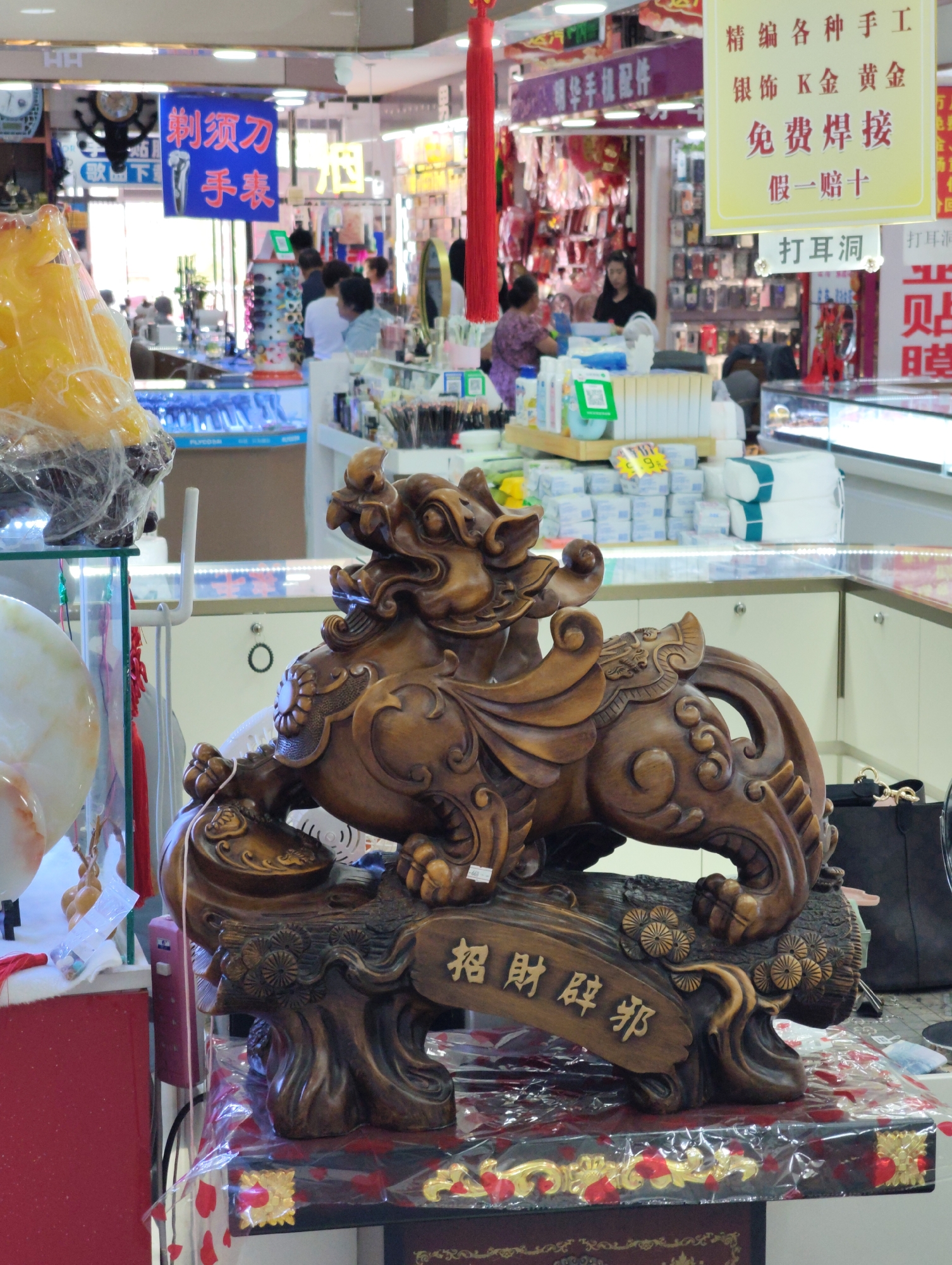 The image depicts a market stall in Lindian, China, likely a jewelry or accessory shop. In the foreground, a wooden carving of a mythical creature, possibly a "Pixiu," sits on a display. The carving is intricately detailed and appears to be a symbol of wealth and protection, with Chinese characters meaning "Attract wealth and ward off evil" inscribed on a wooden plaque at its base. Behind the carving, various merchandise is visible. To the left, there's a glass display case with what appears to be jewelry or decorative items, partially obscured by a large yellow translucent object wrapped in plastic. Further back, shelves are stocked with smaller items, possibly phone accessories or other trinkets. Several individuals are present in the background, engaged in shopping or working at the stall. One person is seen behind a counter, and others are browsing the merchandise. The lighting suggests an indoor environment, typical of a market. Signs in Chinese are visible, indicating the nature of the products or services offered.