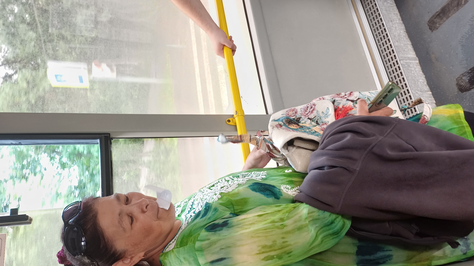 An adult woman with dark hair pulled back and dark sunglasses perched on her head is standing on a bus in Izhevsk, Russia. She is wearing a bright green, tie-dye-style garment with white embroidery around the neckline. A white paper or receipt is held between her lips. She is holding a patterned bag and a dark gray hooded jacket over her left arm. Her right hand is reaching for a smartphone. To her right, another person's hand is visible, holding onto a yellow horizontal handrail on the bus. Outside the bus window, trees and a blurred street sign are visible. The interior of the bus appears to be modern, with a metal handrail and a grated floor vent.
