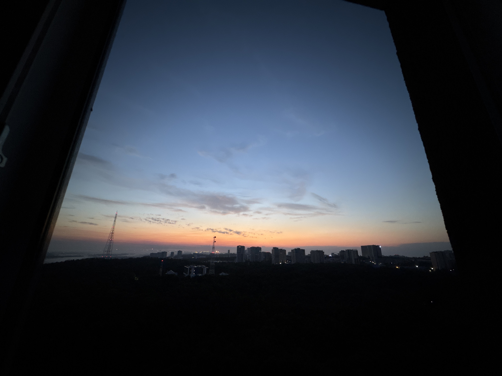 This image captures a twilight view of Ufa, Russia, as seen through a window. The sky transitions from a deep blue at the top to a warm orange and pink hue near the horizon, indicative of either sunrise or sunset. Scattered clouds are visible in the upper part of the sky.
In the middle ground, a silhouetted cityscape stretches across the frame. Prominent structures include several tall buildings, some appearing to be apartment complexes or office buildings. A radio tower, illuminated with red lights, stands out on the left side of the cityscape, piercing the dusky sky. The lower portion of the image is dominated by dark silhouettes of trees and possibly lower-rise buildings, suggesting a green belt or suburban area within the city.
The overall impression is one of urban tranquility at the cusp of day or night. The darkness of the foreground elements, framed by what appears to be the dark interior of a room or structure, emphasizes the vastness of the sky and the distant urban panorama. No people are visible in the image. The scene is static and devoid of any discernible activity.