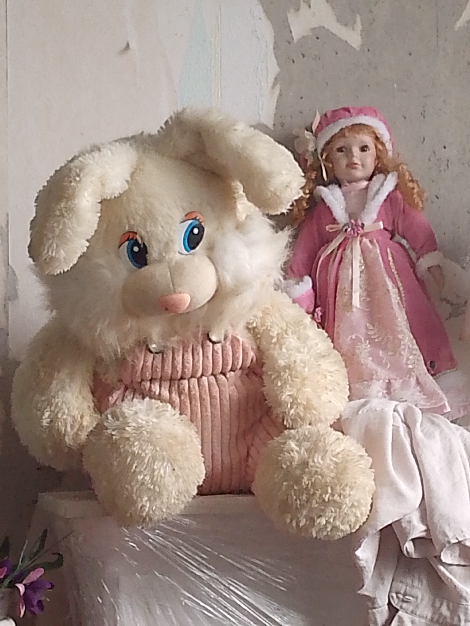 The image shows two dolls in what appears to be an indoor setting. On the left is a large, cream-colored plush bunny with blue eyes and a pink nose, wearing pink overalls. Its right arm is bent, and its left arm is resting on its lap. To the right of the bunny is a porcelain doll with blonde hair, wearing a pink hat and a matching pink dress with white trim. The doll is looking forward with a neutral expression. The background consists of a textured, light-colored wall, possibly wallpapered, with some peeling or damage. In the foreground, to the right of the bunny, is a pile of light-colored fabric, possibly clothing or blankets. To the far left, a few purple and pink flowers are visible, with green stems. The overall impression is of a still-life arrangement of toys. There is no indication of the city of Magadan, Russia, in the image.