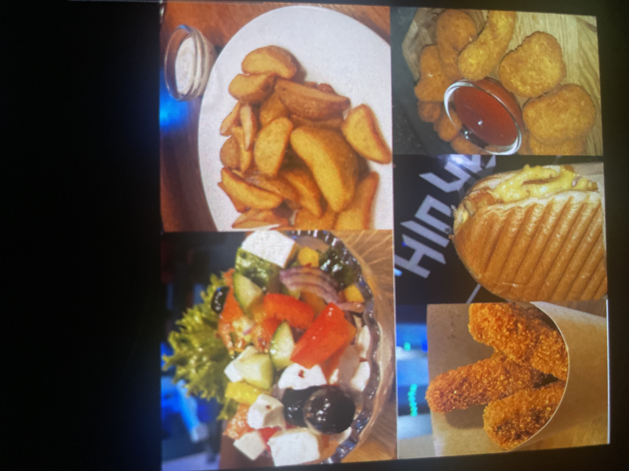 The image displays a collage of various food items. In the top left is a bowl of Greek salad with feta cheese, cucumbers, tomatoes, olives, and lettuce. To its right is a plate of golden potato wedges served with a small bowl of white sauce. The bottom left corner features a paper cone filled with three breaded chicken fingers. Next to it is a grilled sandwich. The bottom right shows a wooden board with chicken nuggets and a small dish of ketchup. The background of the bottom section features text that appears to be "HI N U" in large white letters. There is no indication of the location being in Paczkowo, Poland.