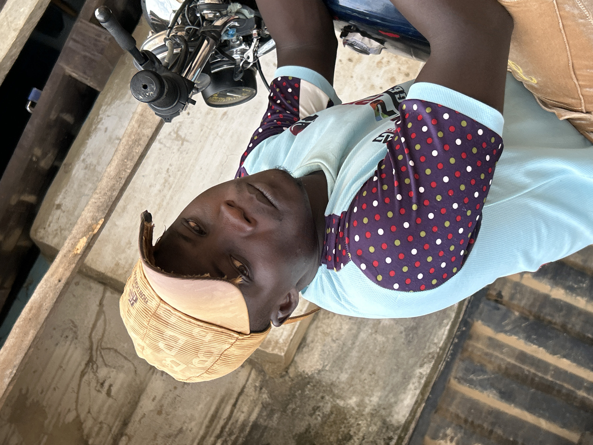A young Black man with dark skin, wearing a light blue t-shirt with a red, green, and white polka dot pattern on the sleeves and a tan corduroy baseball cap, is seated on a motorcycle in what appears to be an outdoor setting. He is looking towards the camera with his head tilted slightly. The background consists of a concrete wall and wooden beams. The scene is likely located in Ihiagwa, Nigeria, given the context.