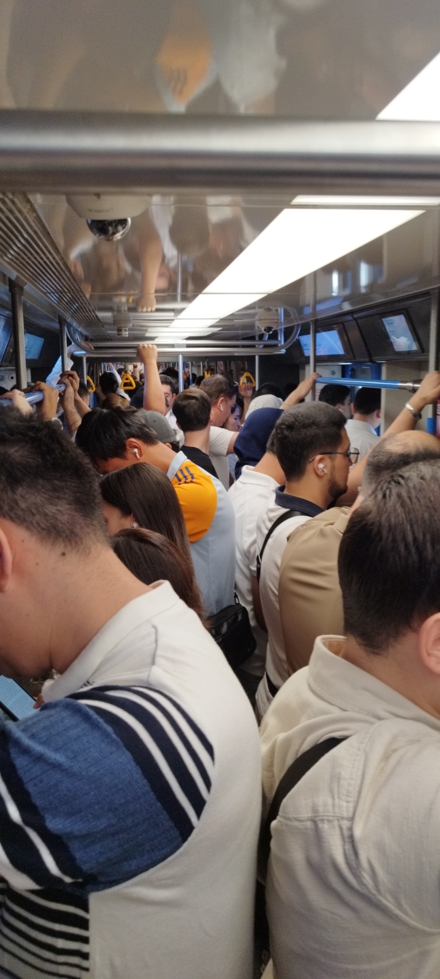 The image shows the interior of a crowded train car. People are standing and holding onto poles and straps for support. Some are looking at their phones, while others are engaged in conversation. The train car is brightly lit with overhead fluorescent lights and has large windows along the sides. The overall impression is one of a busy commute.