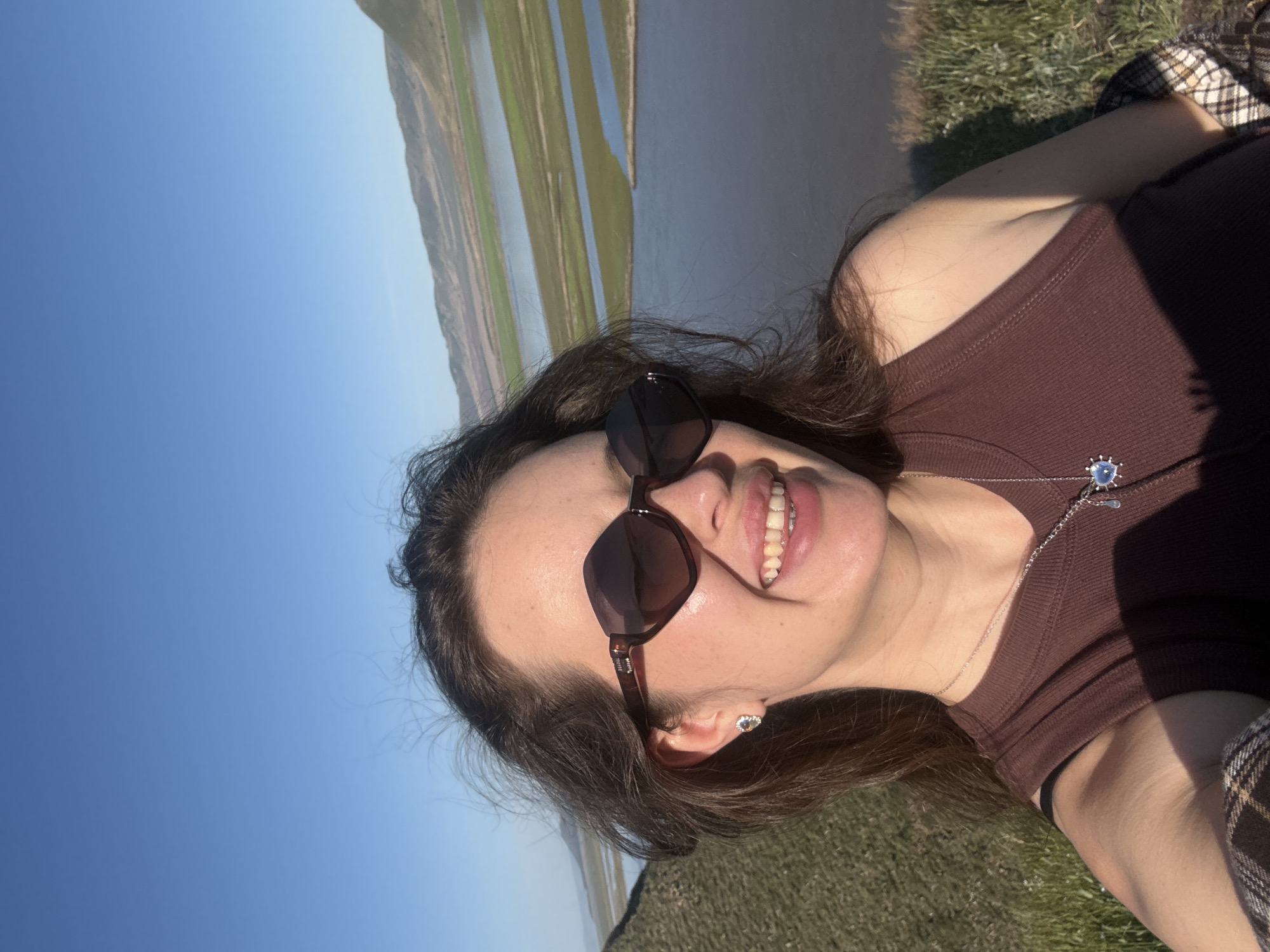 A young woman is depicted in a close-up, horizontally oriented shot, appearing to be lying down outdoors on a sunny day. She is smiling broadly, revealing her teeth, and wearing dark sunglasses that obscure her eyes. Her dark hair frames her face, and she has a small, light-colored stud earring in her visible ear. Around her neck is a thin silver chain necklace with a distinctive light blue and white circular pendant, featuring a silver outline or detailing. She is wearing a dark brown, ribbed tank top or camisole, and a segment of brown plaid fabric is visible on her right side, possibly a blanket or skirt.
The background reveals a vast, clear blue sky to her left. To her right, a wide body of water, likely a river or lake, stretches into the distance, reflecting the sky and suggesting calm conditions. Beyond the water, distant hills or landforms with some green vegetation are visible. The immediate foreground around her consists of green grass, indicating she is relaxing on a grassy bank or field. The bright sunlight creates clear shadows, suggesting a vibrant daytime setting with pleasant weather. The overall impression is one of contentment and relaxation in a natural, open landscape. No visible text is present in the image.