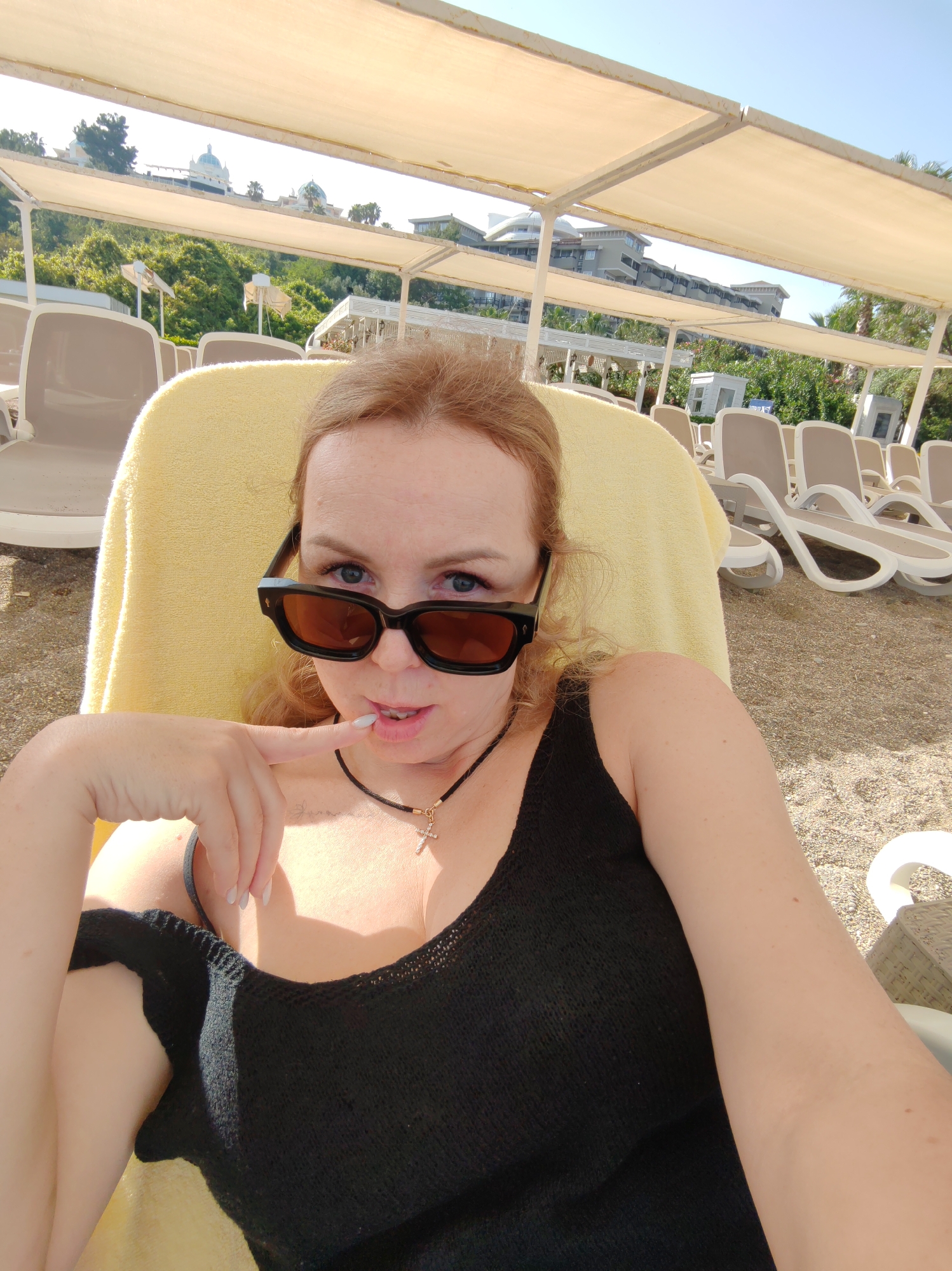 A light-skinned woman is posing for a selfie while sitting in a beach chair. She is wearing a black tank top and dark sunglasses. Her hair is reddish-blonde. She is resting her index finger on her lower lip. She is wearing a necklace with a cross pendant. In the background, beach chairs are visible, along with awnings that provide shade, and a building on a hill. This is likely a beach in Alanya, Turkey. The terrain appears to be gravelly sand.