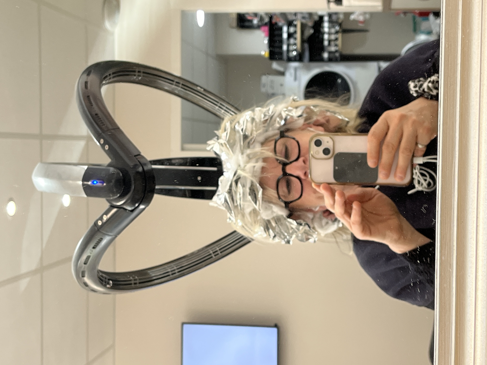 A woman with silver foil in her hair, possibly undergoing a hair treatment or coloring process, is reflected in a mirror. She is wearing glasses and holding a smartphone, seemingly taking a selfie. A large, black hair dryer or styling device is positioned over her head. The reflection reveals a salon setting with equipment and products visible in the background. A television screen is also reflected in the mirror. The location is assumed to be a hair salon in Montrond-les-Bains, France.