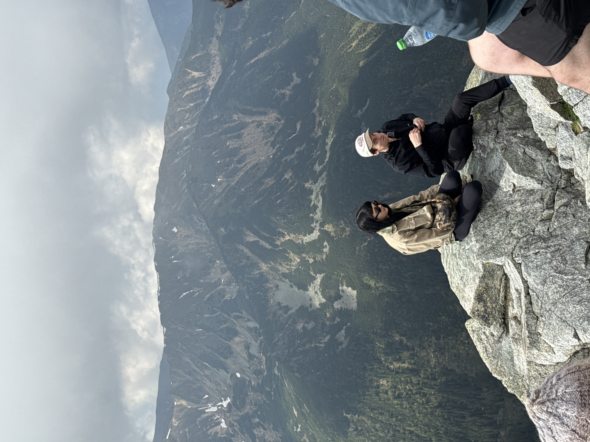 The image, captured in Zakopane, Poland, depicts a group of people resting on a rocky mountain ledge, looking out over a vast mountainous valley. The photograph is oriented vertically, showing the steep drop-off from the cliff edge.
In the upper right, partially cropped, a person wearing a dark top and black shorts holds a plastic water bottle with "Żywiec Zdrój" visibly printed on it. Below them and slightly to the left, another person wears a white baseball cap, a black jacket, and black pants, appearing to gaze at the panoramic view. Further in the foreground, seated directly on the rock edge, is a person wearing sunglasses and a tan-colored jacket (possibly with a subtle camouflage pattern) over dark leggings. A fourth person's knitted clothing is visible in the very bottom right corner, suggesting they are also part of the group.
The people appear relaxed, enjoying the expansive mountain scenery from their high altitude. The valley below is covered in dense evergreen forests with scattered patches of lighter rock and some residual snow. In the background, distant mountain ranges fade into a hazy, cloudy sky. The weather is overcast, and the scene is illuminated by natural daylight, suggesting it's daytime. The rugged terrain and dramatic drop-off emphasize the adventurous nature of the mountain setting.