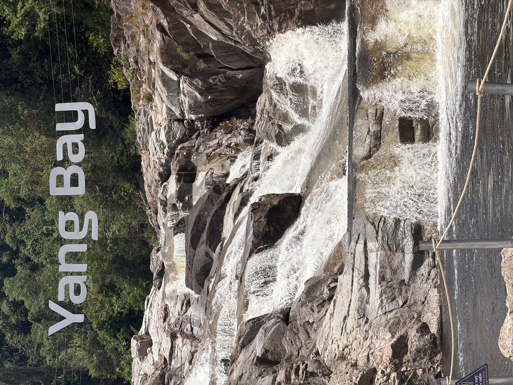 The image depicts Yang Bay Waterfall in Khanh Vinh, Vietnam. Water cascades down a rocky terrain. A sign reads "Yang Bay" vertically on the left. Vegetation is visible to the left of the sign. A metal railing with rope is on the right.
