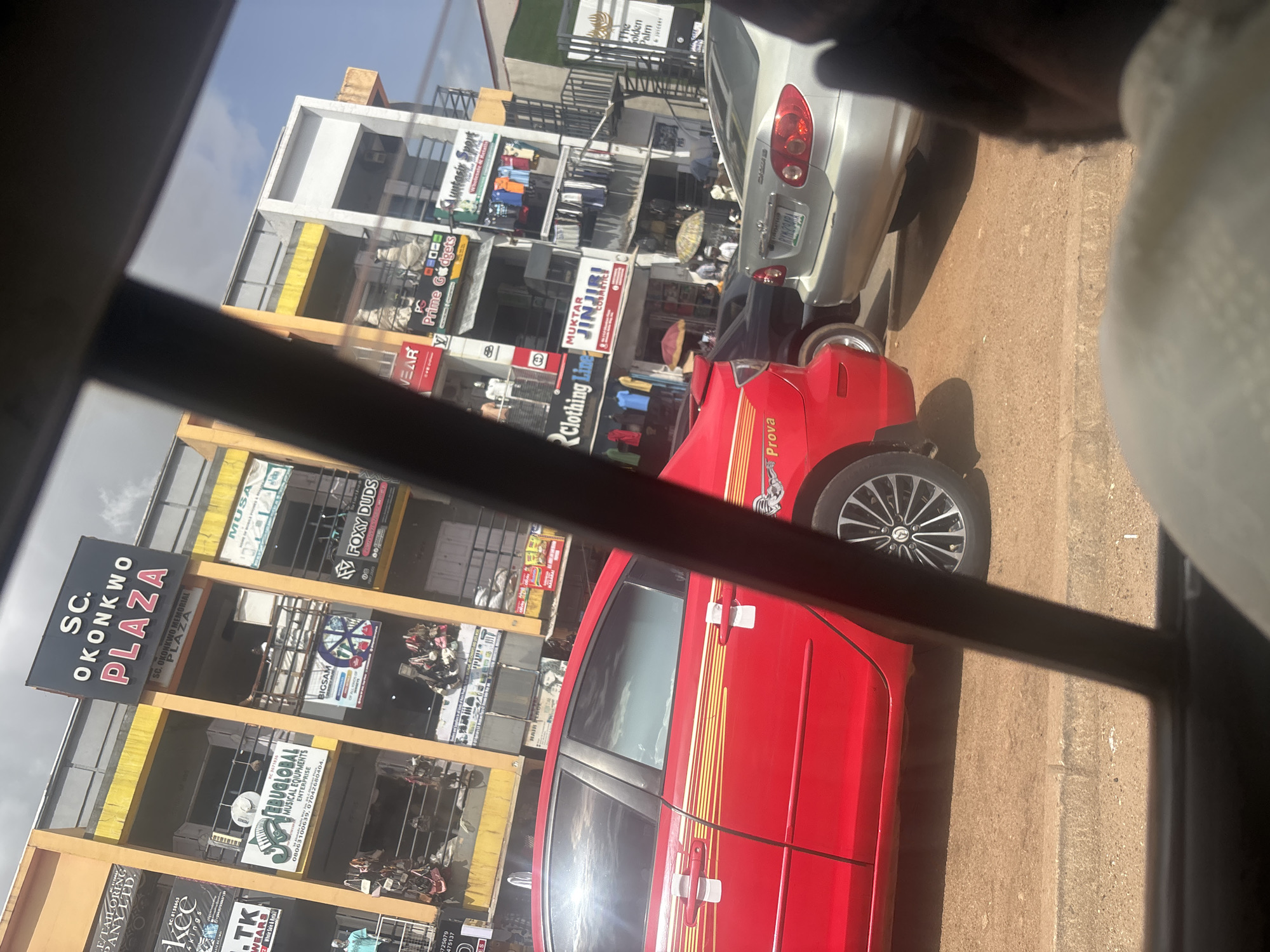 The image shows a street scene in Jos, Nigeria, with a focus on a multi-story commercial building labeled "SC. OKONKWO PLAZA". The building has several shops on different levels, with signs for various businesses. In the foreground, a red car is parked on the street, and a silver car is visible behind it. The street appears to be paved and there are some pedestrians in the background, some of whom are holding umbrellas. The sky is cloudy, and the overall impression is of a busy urban environment.