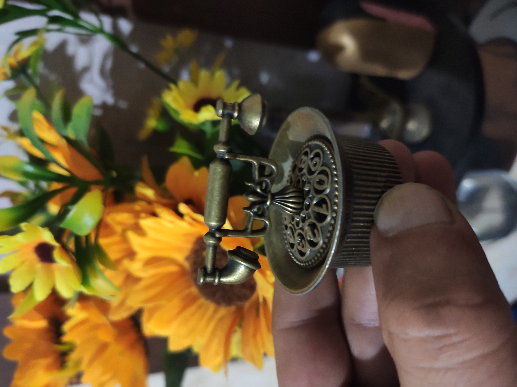 A close-up shot shows a person's hand holding a miniature antique brass telephone. The telephone is ornate with a decorative patterned lid and a ribbed base. In the background, out of focus, is a bouquet of bright yellow sunflowers and greenery. The setting appears to be indoors, with a dark background that suggests a home or indoor display environment. The lighting is soft, highlighting the details of the telephone and the flowers.