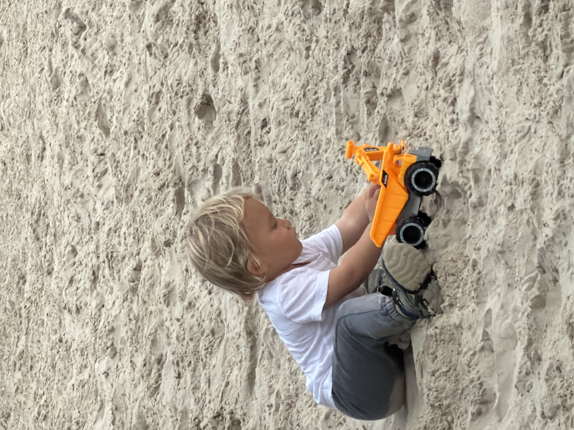 A young child with blond hair and a white t-shirt is positioned against a textured beige surface, likely sand or a sandy wall. The child wears gray pants and sneakers, holding an orange and black toy construction vehicle. The setting appears to be outdoors, possibly near a beach. There's no specific indication of Tel-Aviv, Israel in the image.