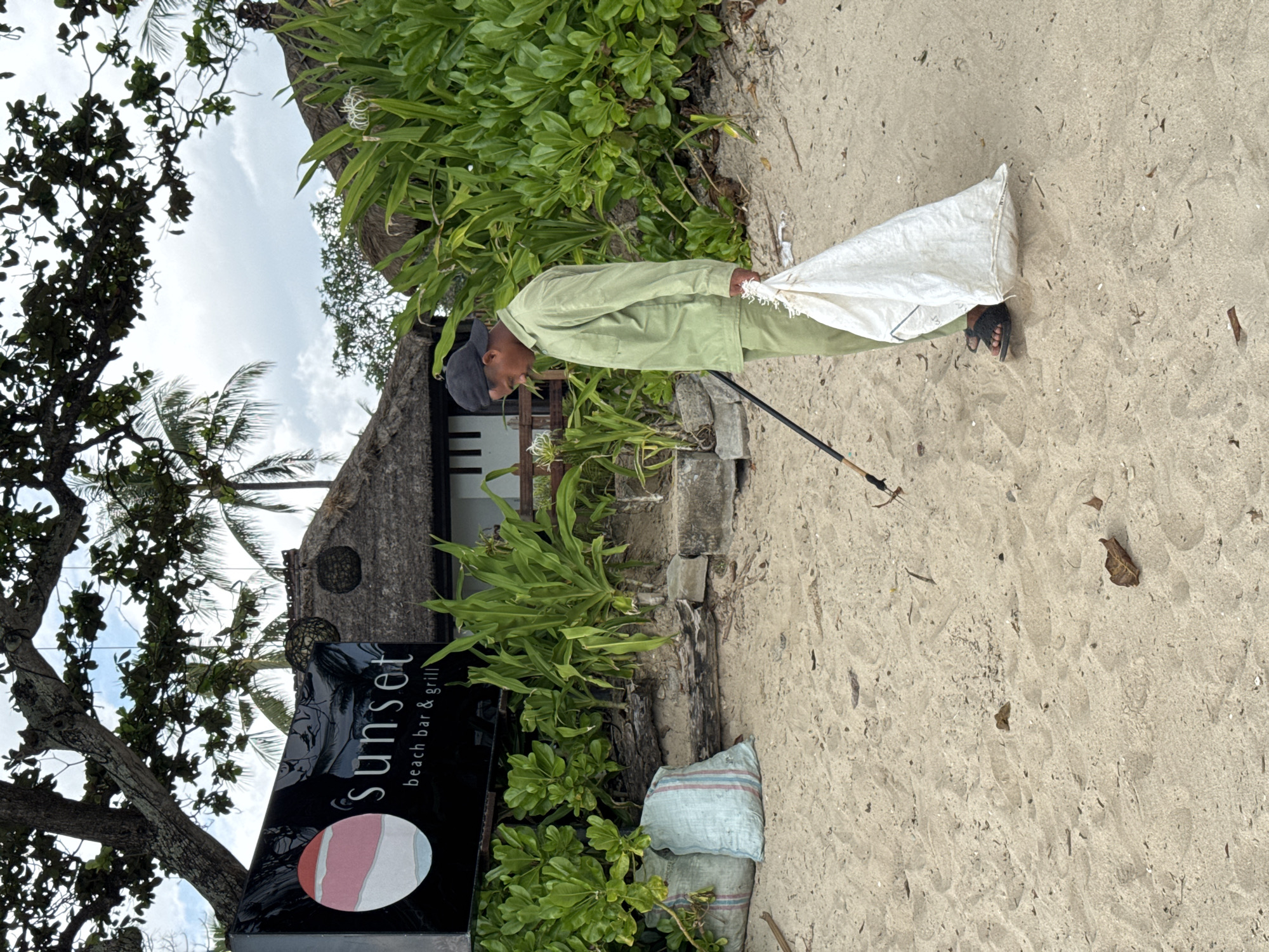 On a sandy beach in Kuta Selatan, Indonesia, a person wearing a light green uniform and a baseball cap is picking up trash with a stick. The person holds a large white bag that appears to contain collected waste. The "Sunset beach bar & grill" sign is visible. A second bag of trash is sitting in the corner, by the side of the restaurant. Tropical foliage is in the background.