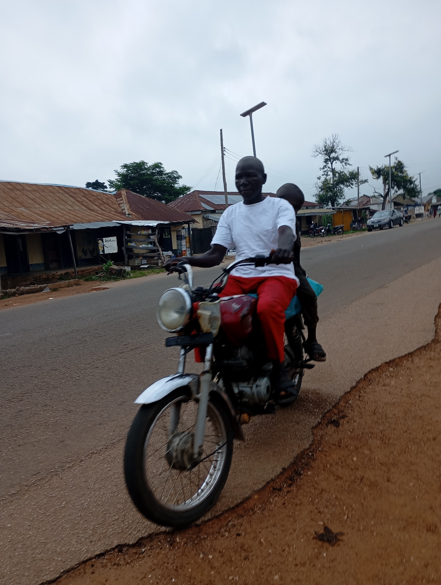 The image depicts a motorcycle carrying two individuals on a paved road in Shendam, Nigeria. The rider, a Black man wearing a white t-shirt and red trousers, is driving. Behind him sits a younger Black individual, wearing a dark shirt and dark trousers. Both individuals appear to be wearing dark sandals. The motorcycle is red and silver. In the background, buildings with corrugated metal roofs line the street, typical of the area. There are also trees and power lines visible. The sky is overcast, suggesting a cloudy day. The road surface is asphalt, with a dirt shoulder on the right side of the image.