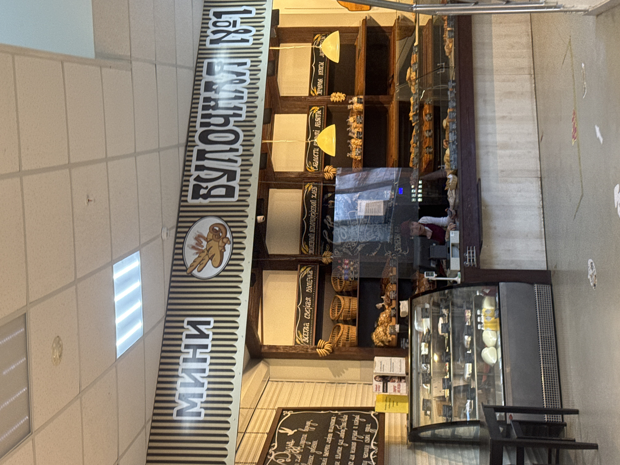 This image shows the interior of a bakery called "Mini Bakery No. 1" in Chaikovsky, Russia. The sign features the name "Mini" vertically in bold, black and white stripes, followed by an image of a gingerbread man or cookie. Adjacent to the sign, the words "БУЛОЧНАЯ №1" are displayed vertically, indicating it is "Bakery No. 1."
Inside the bakery, there are wooden shelves filled with various bread and baked goods. Some shelves have chalkboard signs with white lettering in Russian, possibly describing the items. One sign says "Всегда свежая выпечка" which translates to "Always fresh pastries". Another sign reads "Сладости ручной работы" which means "Handmade sweets". Also a sign reads "ПЕЧЕНЬЯ КЕКС" which means "Cookies Cakes".
A glass display case stands to the right, containing pastries or desserts. Behind the display case, a person is visible, possibly an employee assisting customers. The bakery is well-lit, with tiled ceilings and a neutral-colored wall on the right side. There are black table and chairs in front of the glass display case.