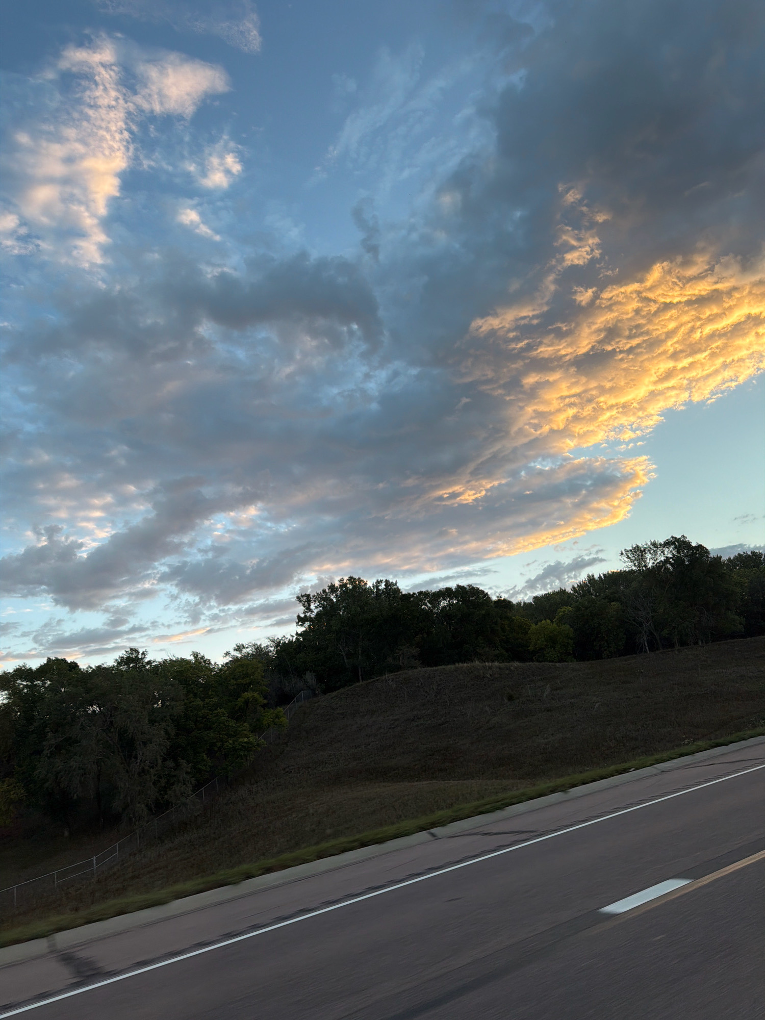 The image shows a highway with a hillside covered in trees and grass. The sky above is filled with clouds, some of which are illuminated by the setting sun, casting a golden glow. The clouds are a mix of white, gray, and golden hues, creating a dramatic and colorful sky. The hillside is covered with dark green trees and dry grass. A chain-link fence runs along the edge of the hillside. The highway is paved and has a white line marking the lanes. The image is taken from a moving vehicle, as indicated by the blurred motion of the road. The overall impression is one of natural beauty and the open road, suggesting a scenic drive.