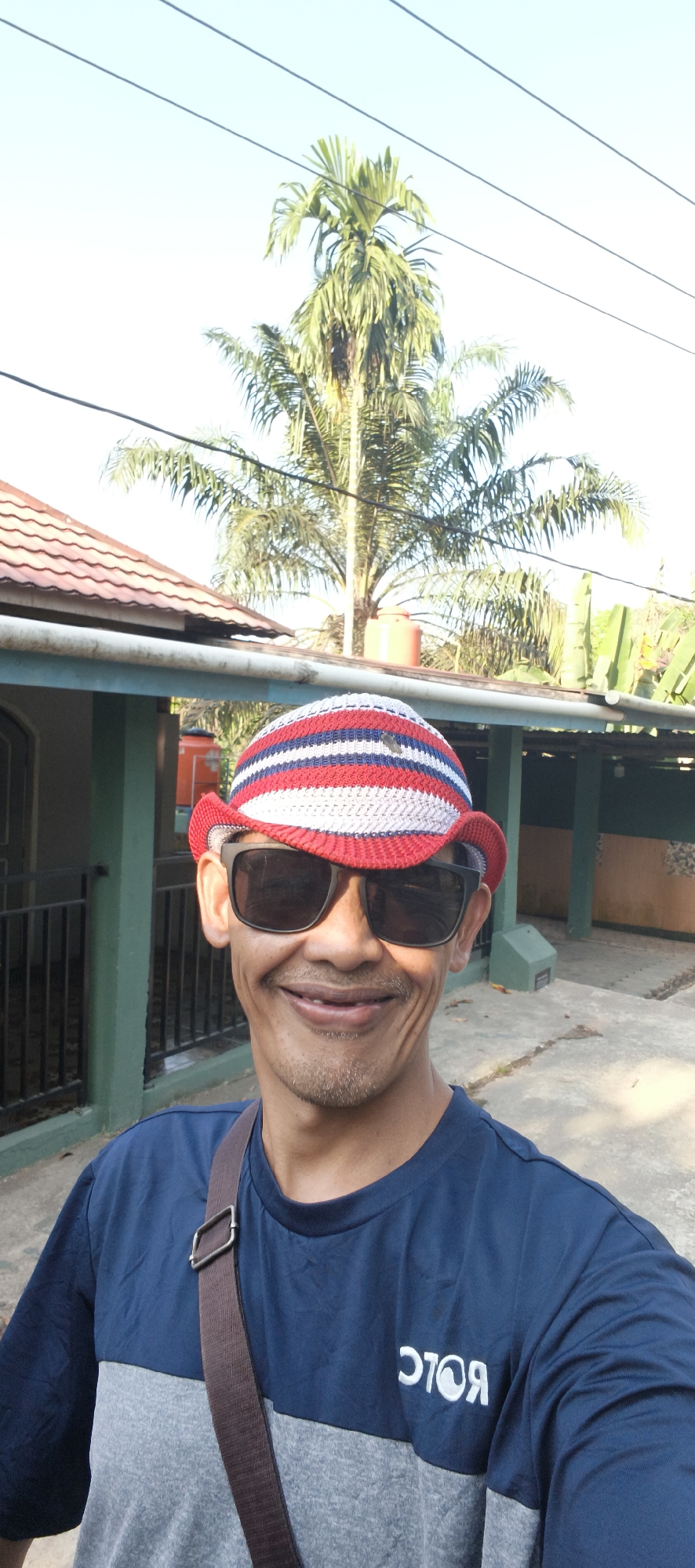 A man wearing a blue and gray t-shirt and a red, white, and blue striped knitted hat stands outdoors, smiling at the camera. He is also wearing black sunglasses. Behind him, a building with a red tile roof and a green exterior is visible. A palm tree and electrical wires are in the background. The ground is concrete. The setting appears to be a residential area in Baru, Indonesia.