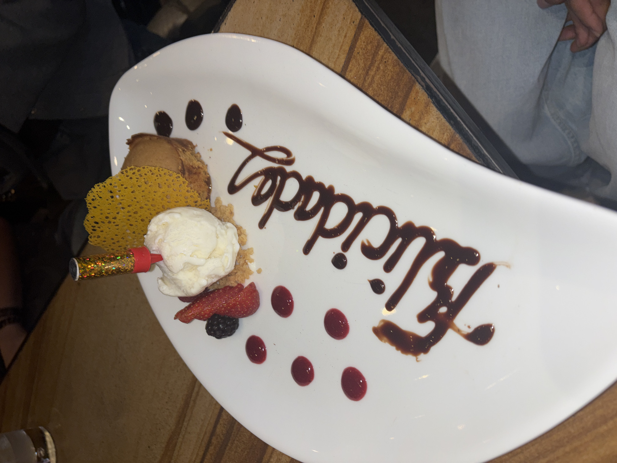 A white plate contains a dessert with ice cream, a piece of pastry, a thin crisp, strawberry slices, and a blackberry. A small firework is embedded in the ice cream. Chocolate syrup is drizzled on the plate, forming the words "Happy Birthday," accompanied by decorative dots of chocolate and red fruit sauce. The plate is placed on a wooden surface, possibly a table, with a person seated nearby, visible by their denim-clad leg. The image does not contain sufficient information to identify the location as Puebla, Mexico, or provide any specifics beyond what is visible in the photograph.