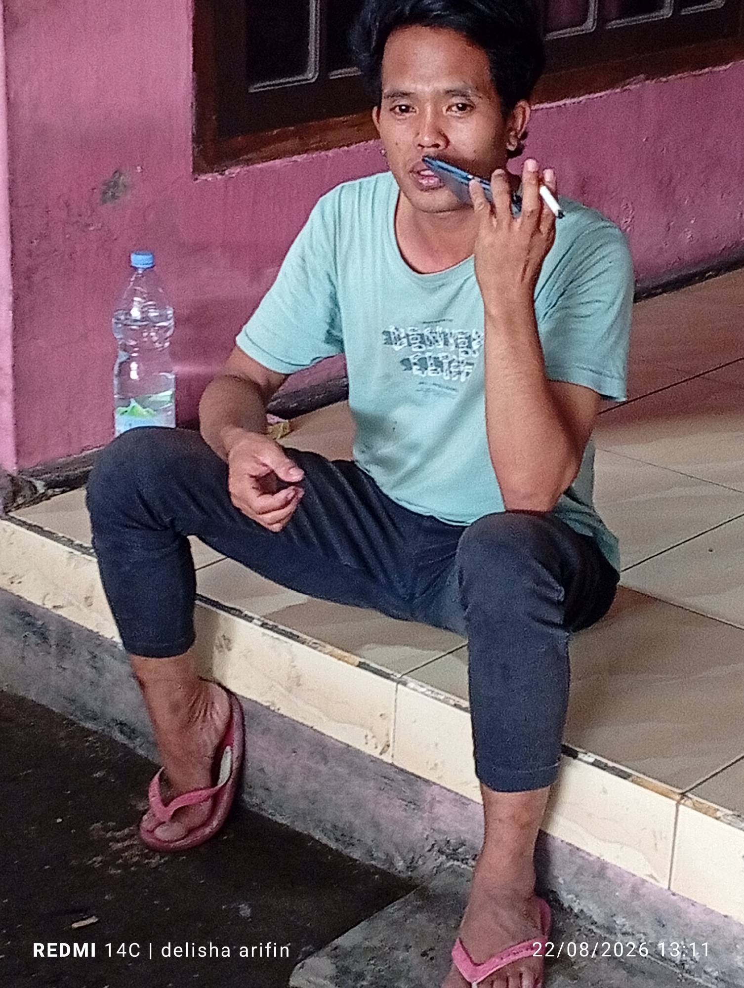 A medium shot shows a man in a teal t-shirt and dark pants sitting on a tiled step. He is holding a white cigarette in his right hand and appears to be speaking into a cell phone with his left hand. A partially full plastic water bottle is to his left, leaning against a pink wall. The scene is outdoors, with the tiled step leading down to a dark, wet surface. The timestamp "22/08/2026 13:11" and the text "REDMI 14C | delisha arifin" are visible in the lower right and left corners of the image respectively. The location is identified as Gambir Kuning, Indonesia.
