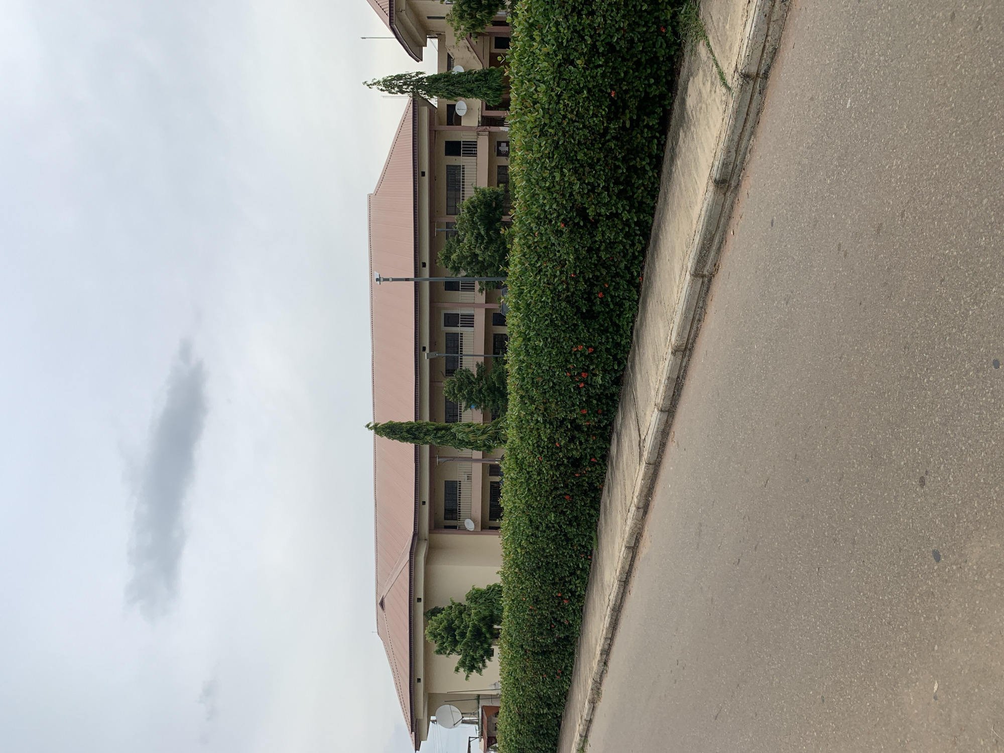 The image depicts a light beige building with a brown metal roof, featuring multiple windows and balconies. A dense, dark green hedge lines the front of the building, with a sidewalk and an asphalt road in the foreground. The sky is overcast with a few scattered clouds. The location is identified as the Federal University Wukari in Wukari, Nigeria.