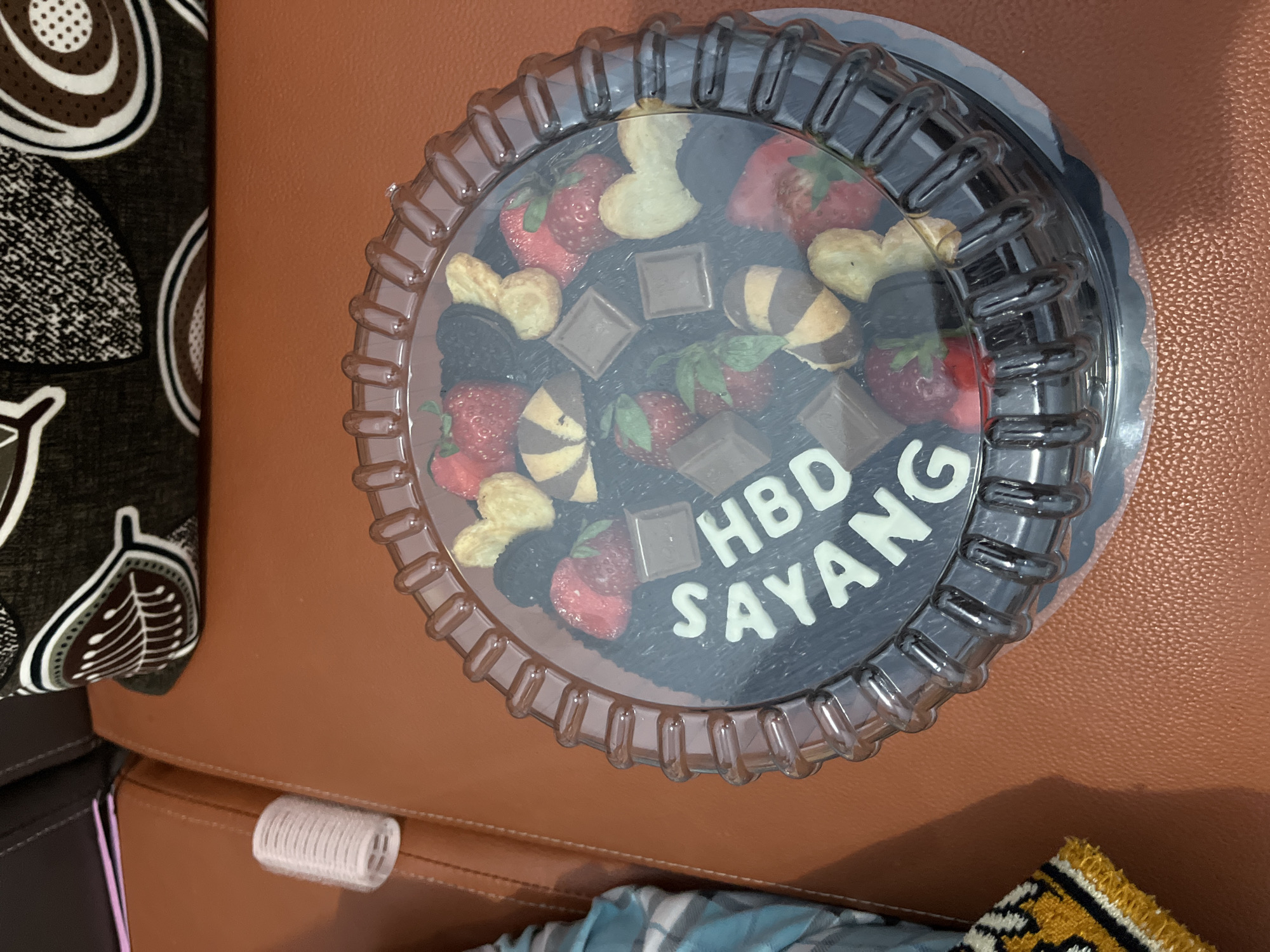 A circular chocolate cake with white icing letters spelling "HBD SAYANG" is displayed in a clear plastic container. The cake is decorated with strawberries, chocolate squares, cookies, and possibly black cherries. The cake is placed on a brown leather surface, likely a couch. In the background, a patterned throw pillow and a pink folded paper are visible. To the left of the cake, a small white cylindrical object, possibly a hair roller, rests on the brown surface. A portion of a blue and white checkered fabric is also visible. The scene is an indoor setting in Jambearum, Indonesia.