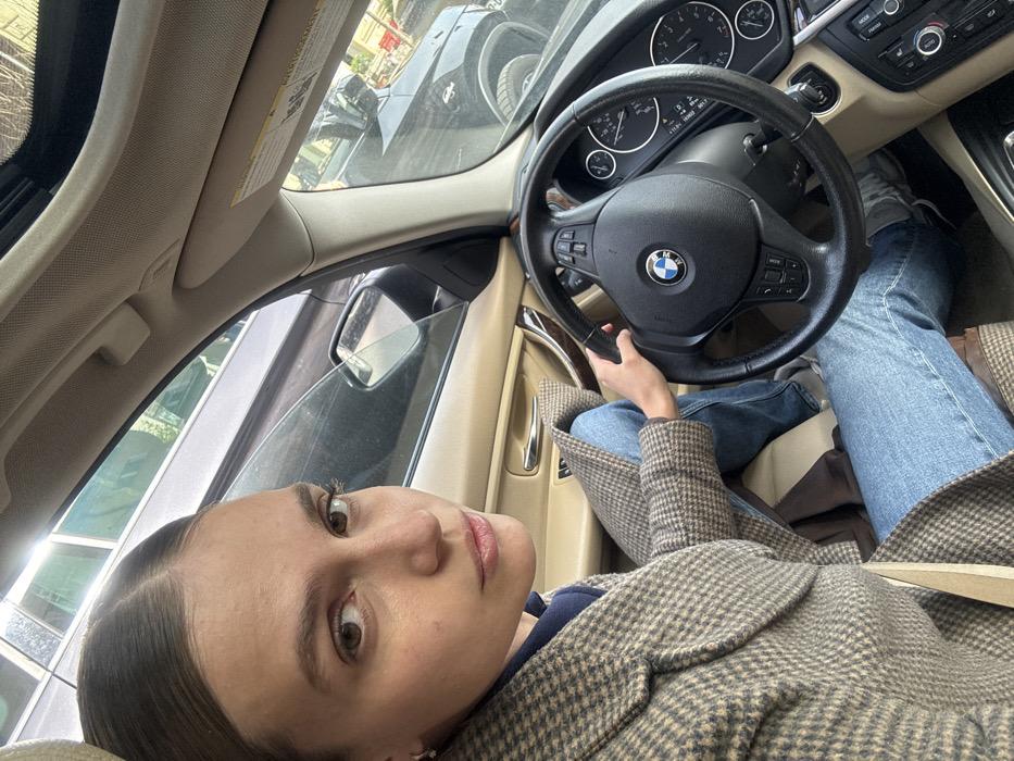 An adult female is depicted seated in the driver's position of a BMW vehicle with a beige interior. She has brown hair pulled back, brown eyes, and maintains direct eye contact with the camera. Her attire consists of a brown and beige houndstooth or plaid coat over a dark blue garment, paired with blue denim jeans. Her left hand rests on the black BMW steering wheel. The external environment, viewed through the vehicle windows, reveals an urban setting with other parked vehicles, including a dark SUV, and reflections of modern buildings. Daylight illumination is present. This scene is located in Tbilisi, Georgia.