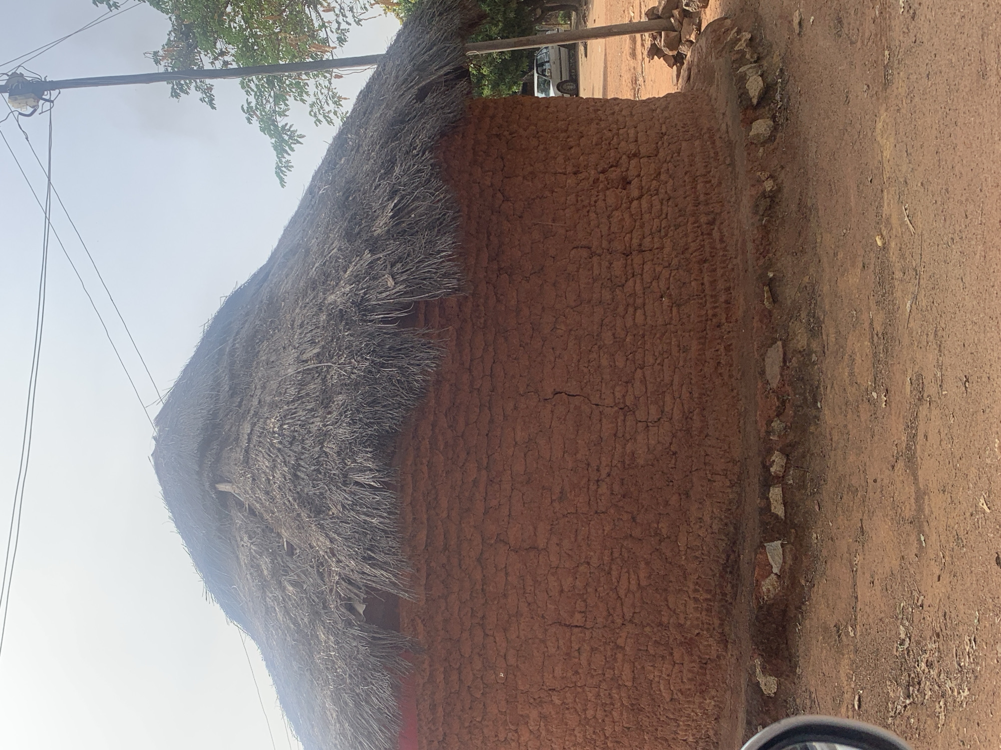 The image shows a mud brick building with a thatched roof. The building is located next to a dirt road. There are trees and telephone poles visible in the background. Based on the provided information, this scene is likely in Jos, Nigeria. There are no people visible in the image.