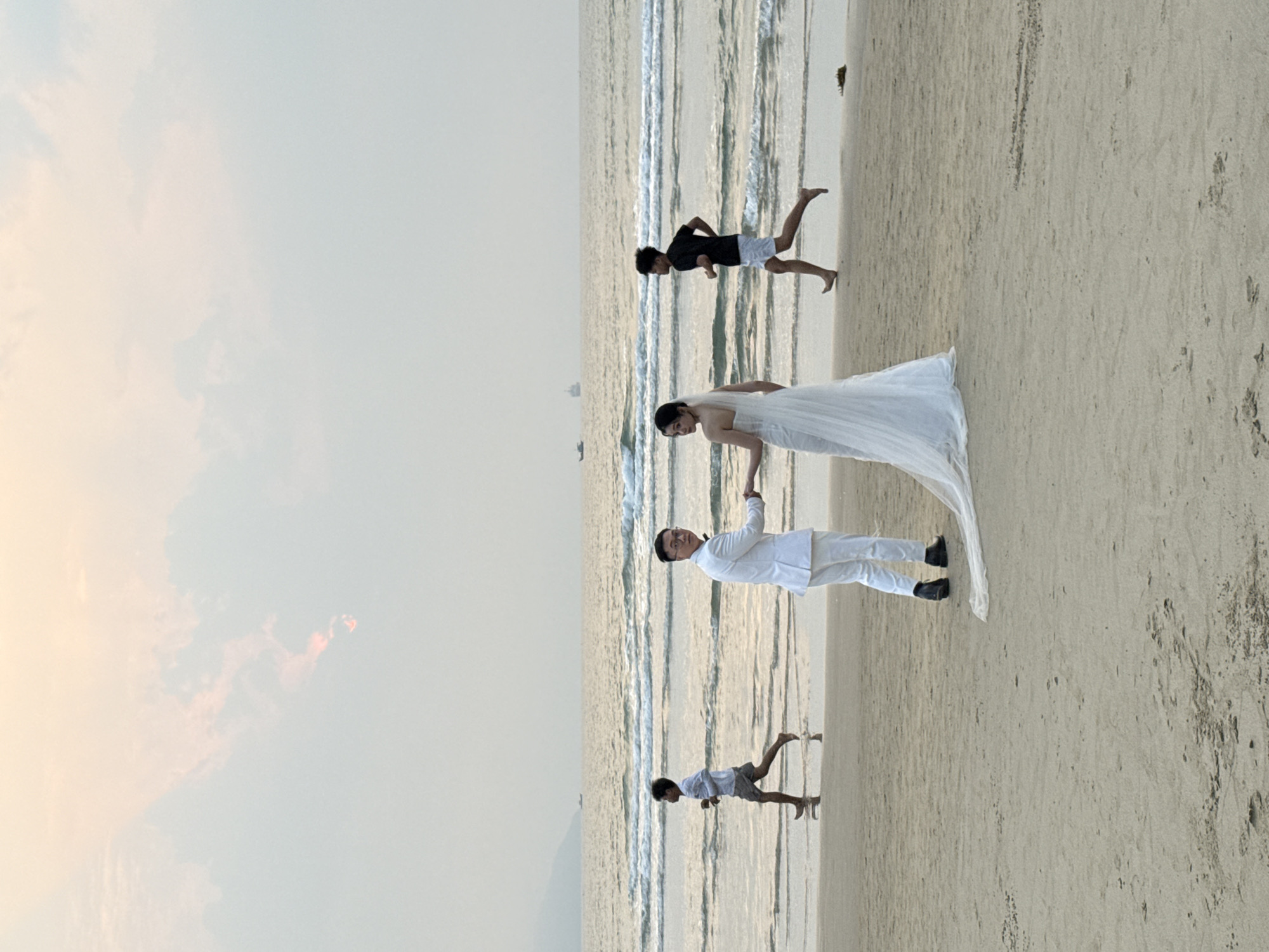 A couple, dressed in white wedding attire, stands on a sandy beach. The bride wears a white wedding dress with a long train, and the groom is in a white suit. Two young people are near the waves. The sky is visible, showing the sunset. The location is likely a beach in Đà Nẵng, Vietnam.