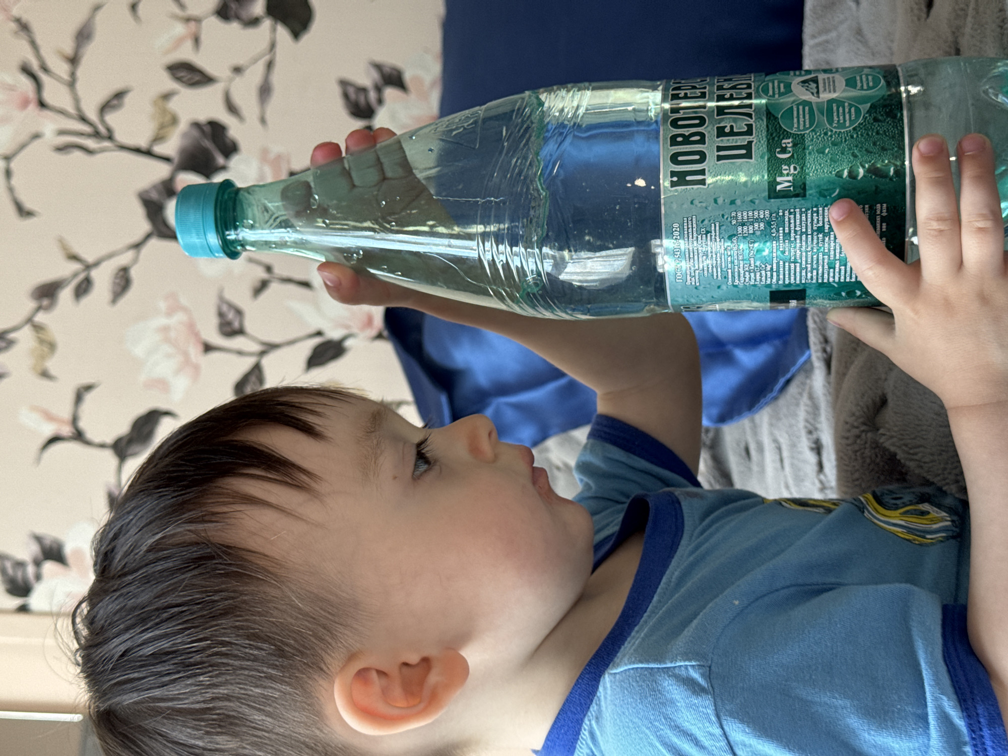 A young child, with dark hair and a blue shirt with dark blue trim, is holding a plastic bottle of "Novoterskaya Celebnaya" mineral water, a brand sourced from the city of Sochi, Russia. The child is tilting the bottle to drink, their mouth open and eyes looking upward towards the bottle. The bottle's label is partially visible, displaying mineral content information in Russian and stating "Mg Ca" prominently. The background includes a floral wallpaper and a blue fabric draped over what appears to be a gray cushion. The image is slightly rotated clockwise.