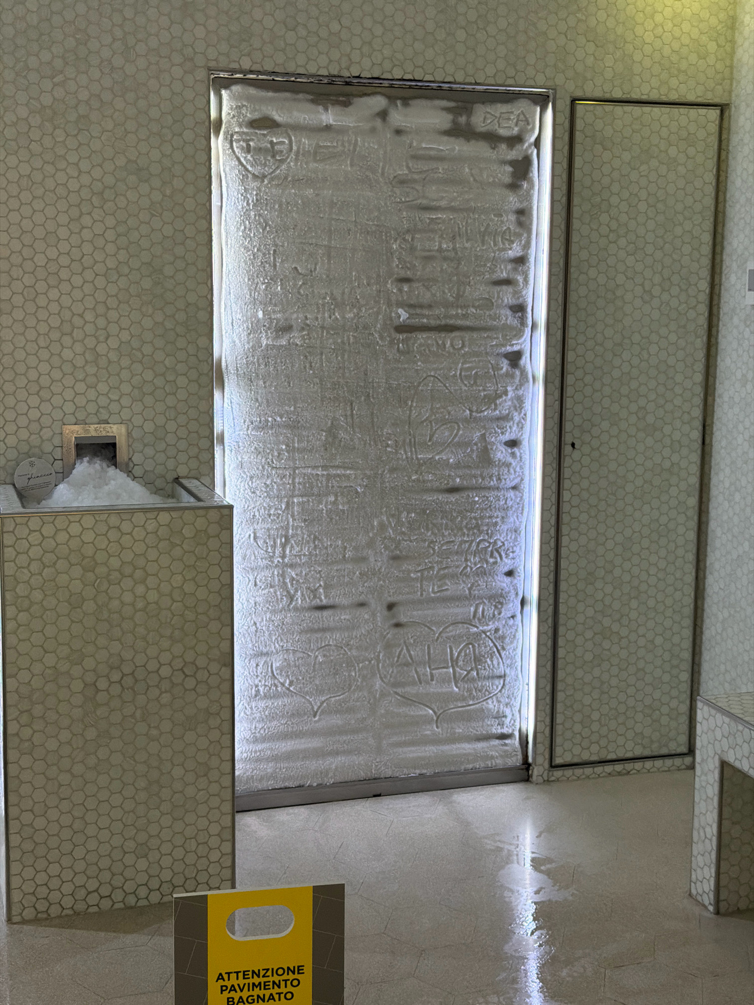 The image depicts a cryotherapy chamber in the Grand Hotel San Pellegrino in San Pellegrino Terme, Italy. The chamber features a frosted glass door with various inscriptions and drawings made in the ice. To the left of the chamber is an ice dispenser. The floor is wet, and a yellow caution sign with "ATTENZIONE PAVIMENTO BAGNATO" (Attention, wet floor) is visible. The walls are tiled with small hexagonal tiles.
