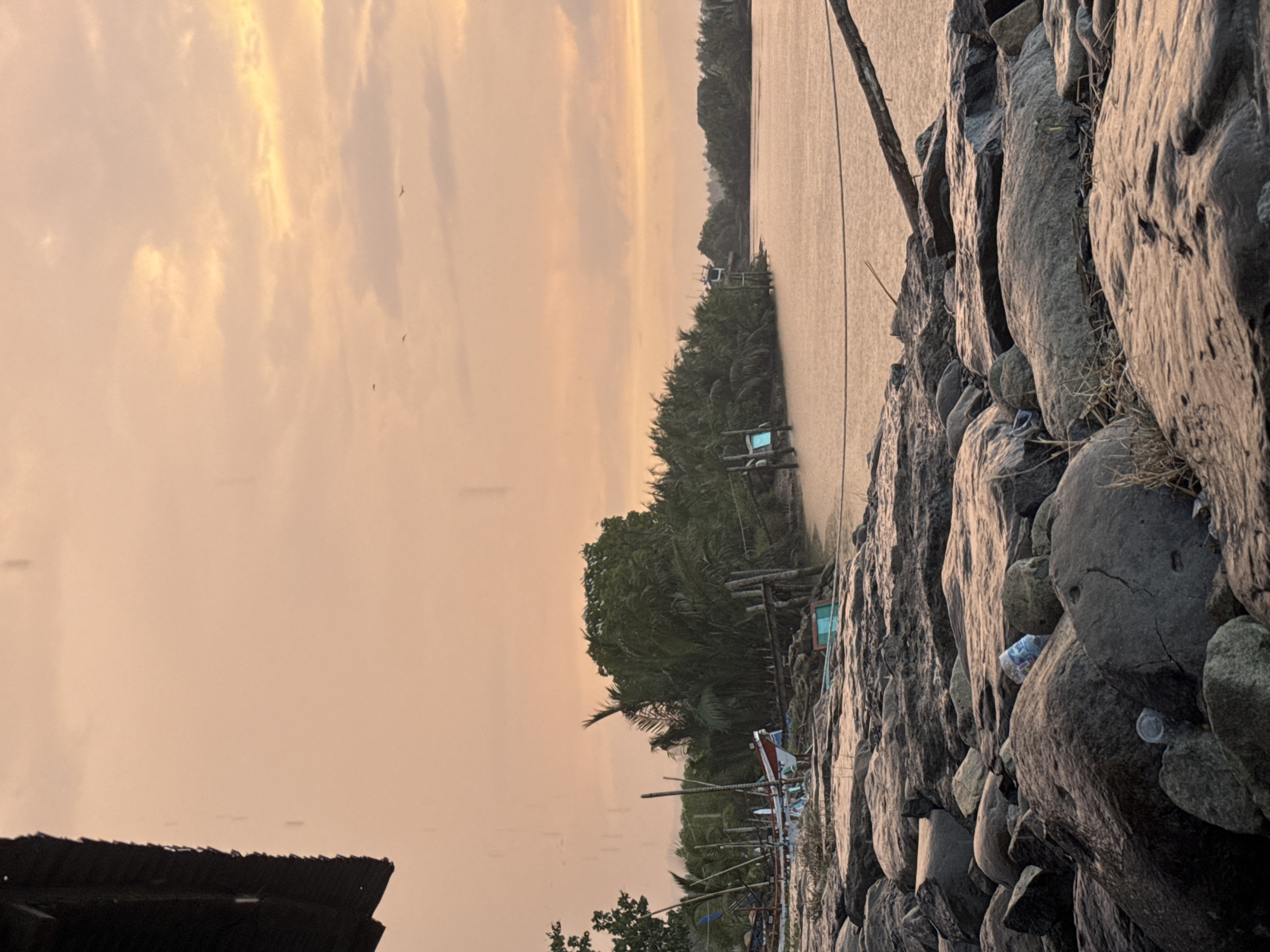 This image depicts a waterfront scene at dusk in Reudeuep, Indonesia. The sky is a muted orange and pink, with scattered clouds and a few distant birds. In the foreground, a rocky shoreline is visible, with some scattered plastic bottles and debris. A portion of a dark, corrugated roof structure juts into the frame from the upper left. Beyond the rocks, a calm body of water stretches towards a lush, green treeline. Several small fishing boats are docked along the shore, some appearing weathered and in disrepair. Wooden poles and ropes are also present, suggesting fishing activity. A few wooden structures, possibly for fishing or observation, stand in the water near the treeline. The overall atmosphere is tranquil, with a hint of human activity through the presence of the boats and structures.