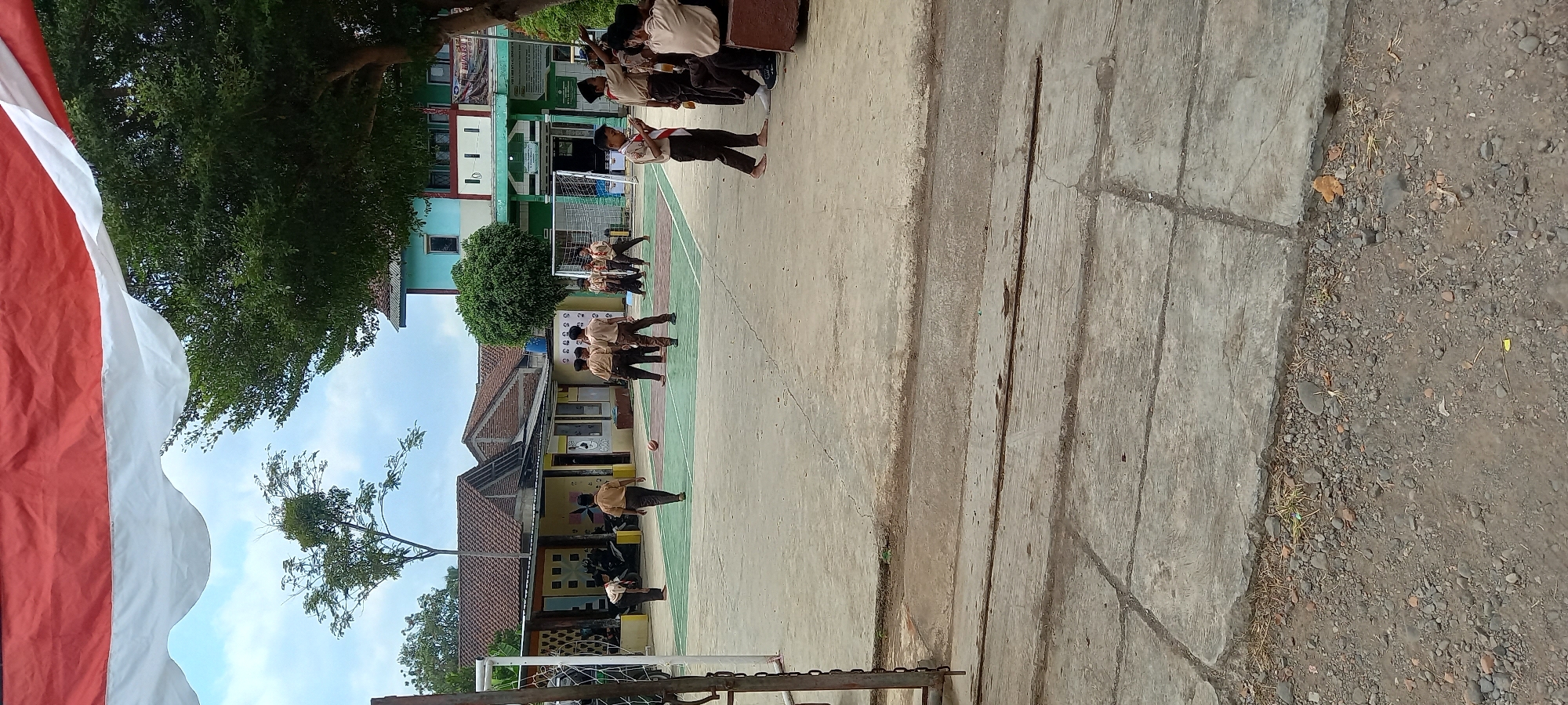 The image depicts an outdoor scene with children in uniform on what appears to be a school campus. In the foreground, a paved area shows cracks and is bordered by gravel. In the midground, several boys wearing brown uniforms are engaged in an activity on a court marked with green and white lines. Some are standing, while others appear to be kicking or observing a red ball. Behind them, a building with a light blue and green facade is visible, featuring multiple windows. A soccer goal is set up on the court. To the left, a white fence is partially visible. Overhead, a red and white draped banner is present. The sky is overcast. The scene is identified as taking place in Cintaratu, Indonesia.