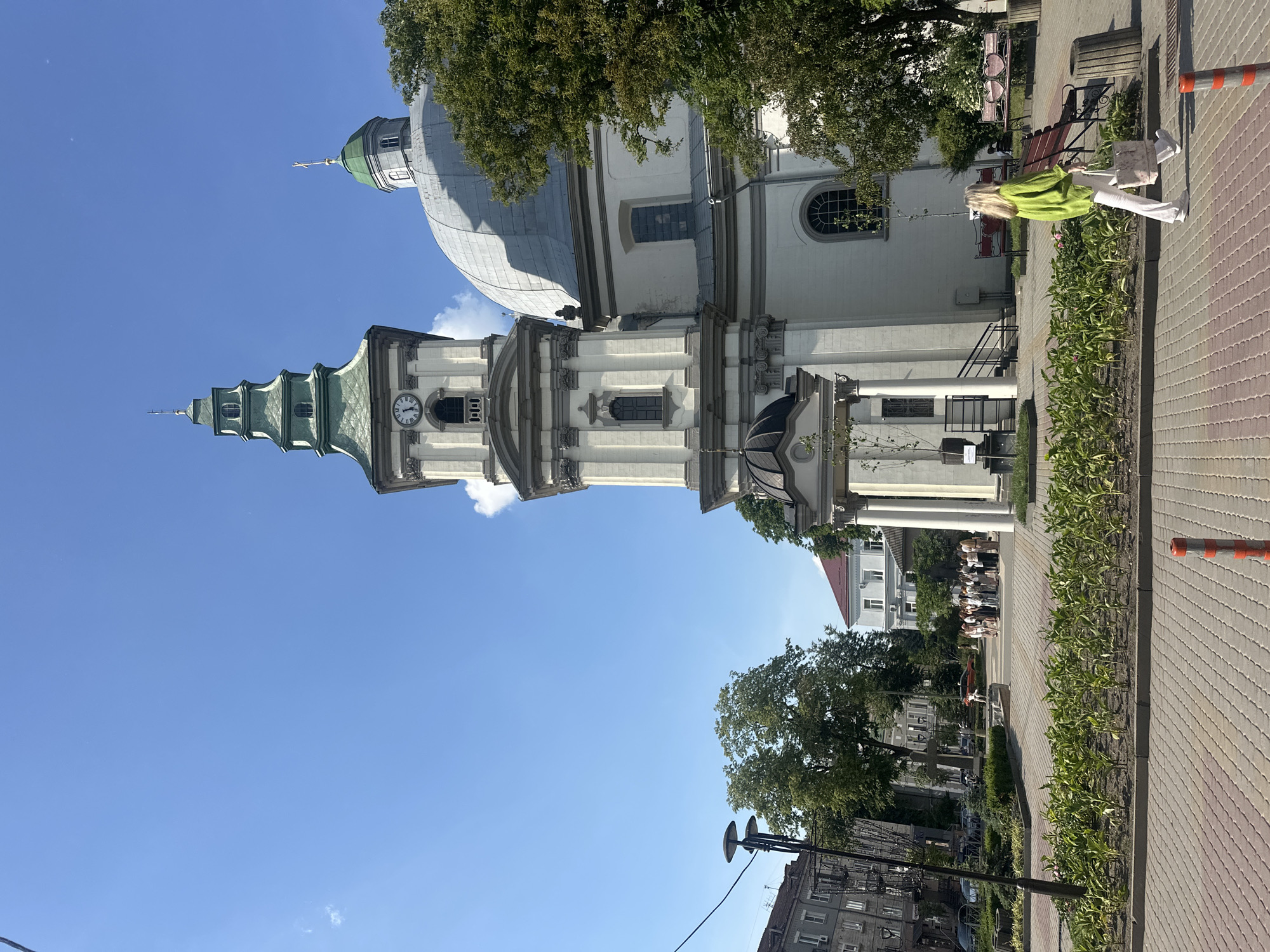 This is a vertical, eye-level, outdoor shot of a church and its surroundings on a sunny day. The church, with its white facade and green-spired tower, dominates the left side of the frame, with a large dome visible behind the tower. A clock face with Roman numerals is prominent on the tower. The sky is clear and blue, with a few wisps of white clouds.
In the foreground, a woman in a bright green sweater and white pants is walking on a paved walkway lined with lush green plants. She is carrying a bag and appears to be heading towards a bench. To the right of the walkway, a park bench with decorative red heart-shaped backs is visible, and further down the path, a trash can and a red and white striped pole with an orange cap are present.
In the background, across the street from the church, several people are gathered, and cars are parked. Trees with full green foliage provide shade in various parts of the scene. The overall atmosphere is peaceful and pleasant, suggesting a typical day in this Ukrainian city. The time of day appears to be late morning or early afternoon due to the bright sunlight and clear sky.