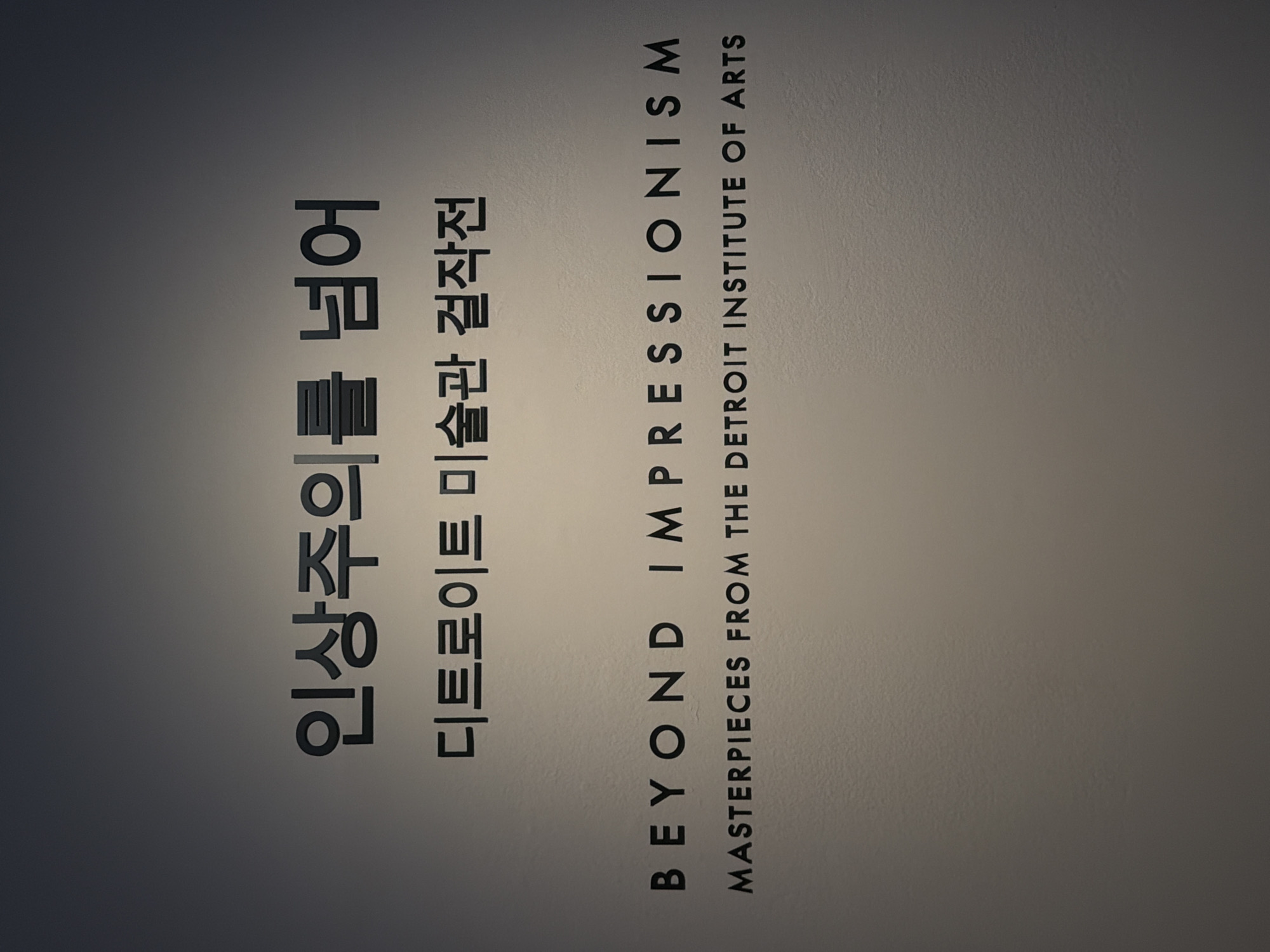 The image shows a wall with text. The text is in Korean and English. The Korean text reads "인상주의를 넘어" and "디트로이트 미술관 걸작전". The English text reads "BEYOND IMPRESSIONISM" and "MASTERPIECES FROM THE DETROIT INSTITUTE OF ARTS". The text is black and appears to be raised from the wall. The wall is a light gray color and is textured. The lighting in the image is dim, with a spotlight shining on the text, creating a gradient effect on the wall. This appears to be an exhibit title, likely from a museum. The location is not specified in the image.