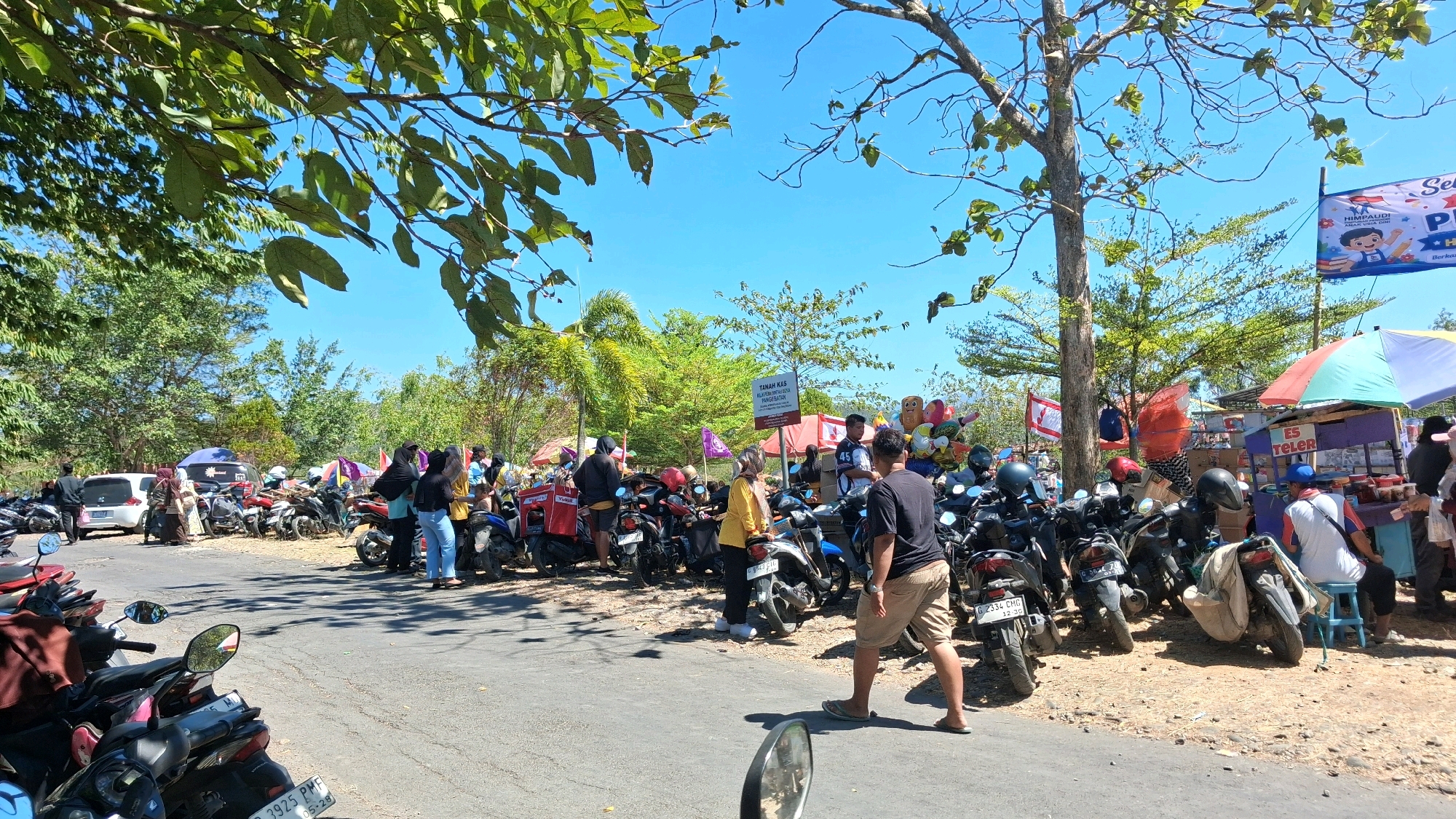 The image depicts a street scene in Parasa, Indonesia, likely a local market or event on a sunny day. A paved road is lined with numerous parked motorcycles. People are scattered throughout the scene, some standing and conversing, others walking.
In the foreground, a motorcycle's side mirror is visible, suggesting the photo is taken from another motorcycle. Further down the road, several individuals are gathered around a white car. To the right of the road, various stalls are set up, with one stall displaying colorful balloons. Another stall has an umbrella and appears to be selling drinks, with "ES TELOR" visible on signage.
A banner with an illustration of a child and text in Indonesian is displayed on a pole, possibly indicating an educational event or advertisement. The environment is characterized by trees and lush green vegetation, with a bright blue sky overhead. The overall atmosphere suggests a vibrant, outdoor gathering.