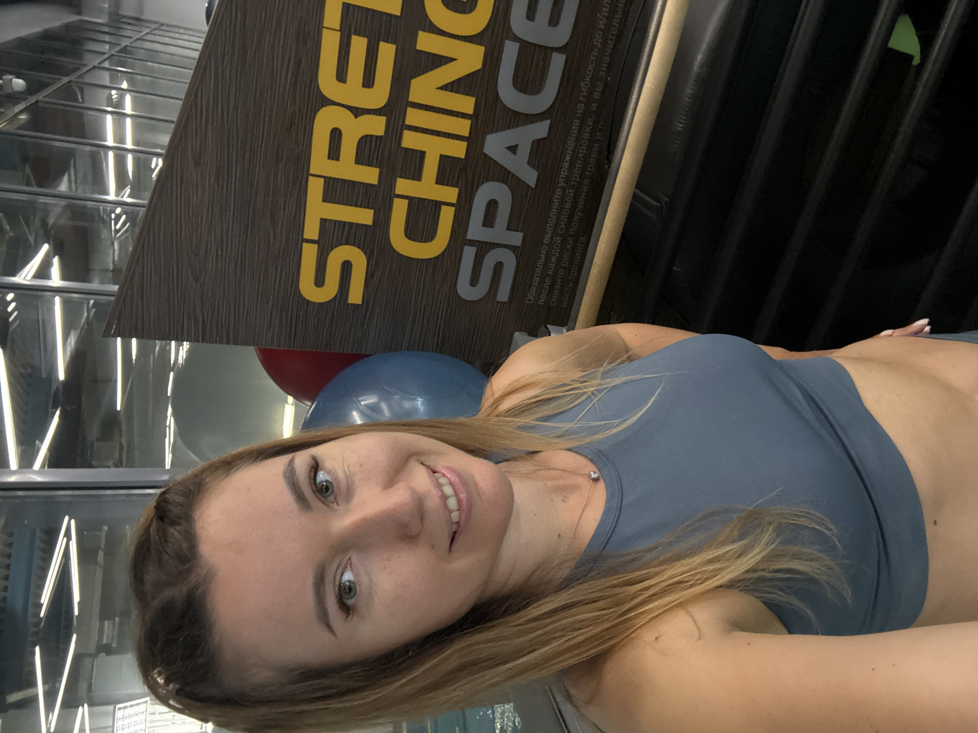 A portrait shot shows a woman in front of a stretching space sign. She is wearing a blue sports top, and there are blue and red exercise balls behind her. The sign reads "STRETCHING SPACE" in bold letters, and there's some Russian text about flexibility exercises after strength training. The location appears to be in a gym or fitness center.