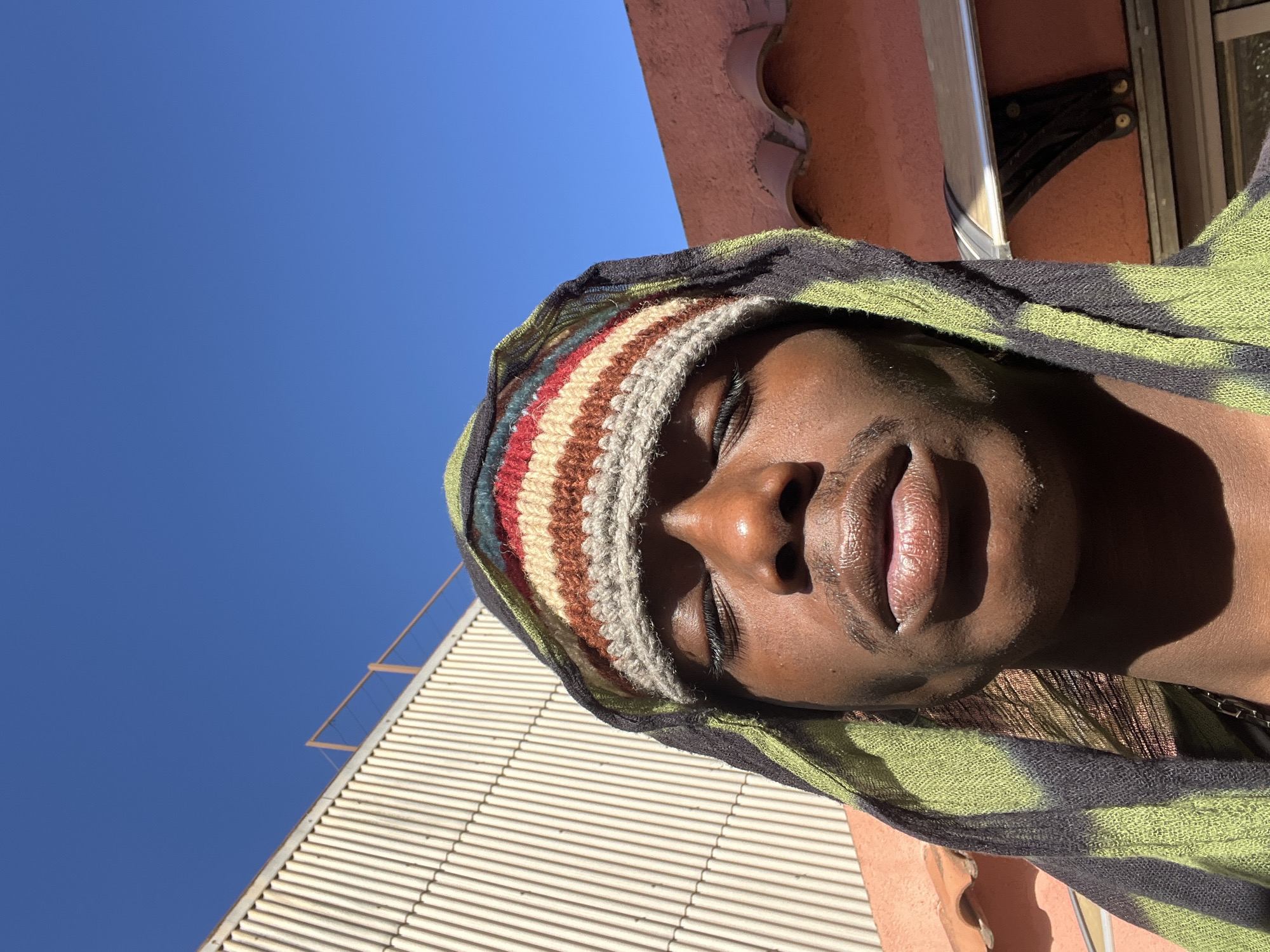A close-up, upward-angled photograph depicts a dark-skinned person wearing a striped knit cap with a green and black striped scarf draped around their head and shoulders. The person's eyes are closed, and they are facing upwards. Behind the person, the clear blue sky is visible. In the lower-left corner, a white building with corrugated metal walls can be seen. To the right, a terracotta-colored building is partially visible, with some architectural details like a window or a protruding section.
The location in the city of Barcelona, Spain cannot be determined from the visual information provided. No identifying architectural landmarks or contextual clues are present to ascertain the specific location.