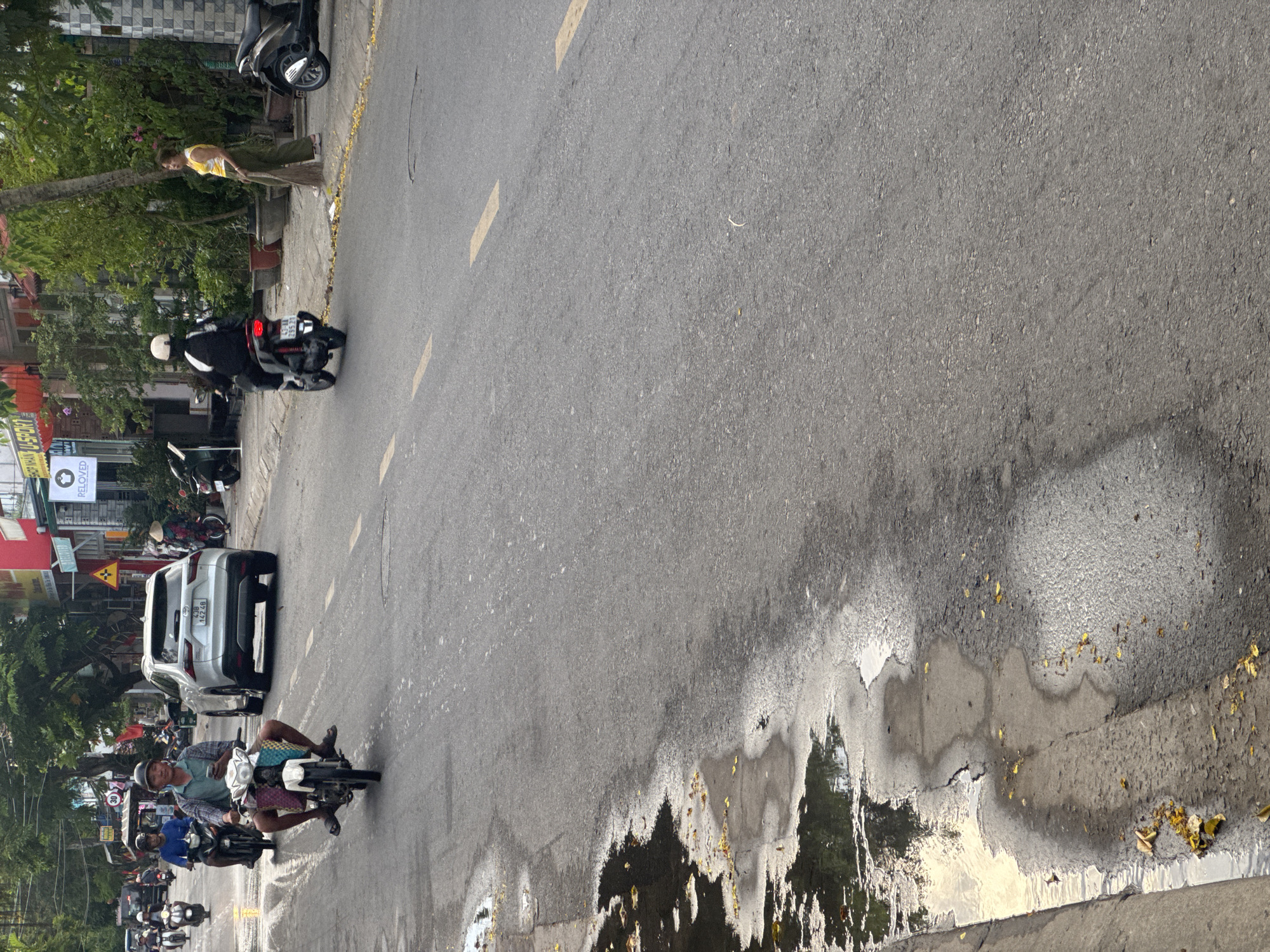 The image shows a street scene in Đà Nẵng, Vietnam. The road is mostly asphalt with some areas showing water puddles and debris along the curb. A woman is sweeping the sidewalk near a tree. Several motorcycles and a silver SUV are present, moving in the same direction. A group of motorbikes are travelling close together. The license plate of the SUV is partially visible. Buildings and storefronts with Vietnamese signage line the other side of the street. The lighting suggests it is daytime. Yellow road markings can be seen.