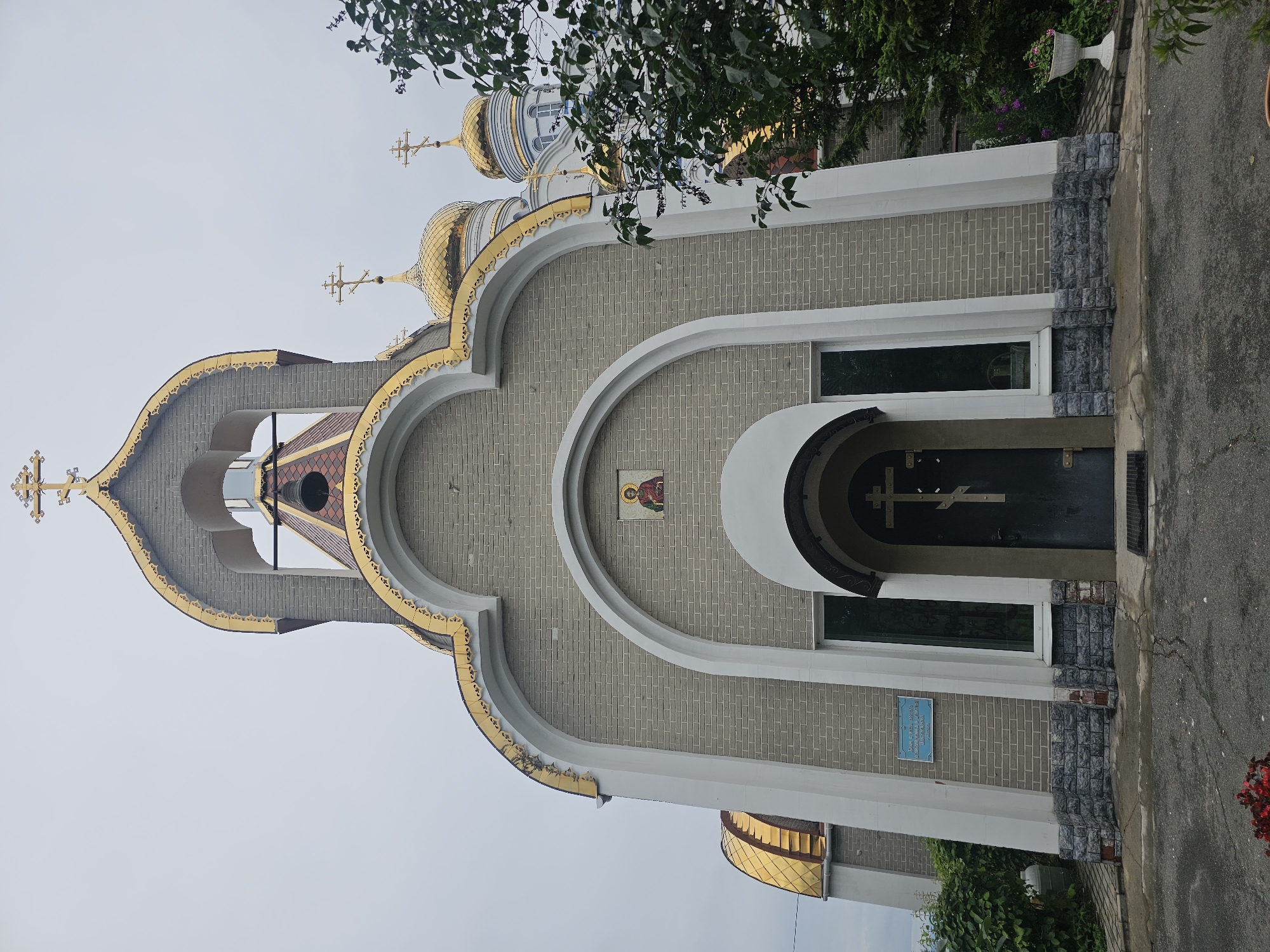 The image shows the exterior of an Orthodox church, likely the Church of the Resurrection of Christ in Nakhodka, Russia. The church has a bell tower with a golden dome and cross, and a gray brick facade. There is a dark colored door with a cross on it, and an icon above the door. The church is surrounded by greenery, including trees and bushes. The sky is overcast.
