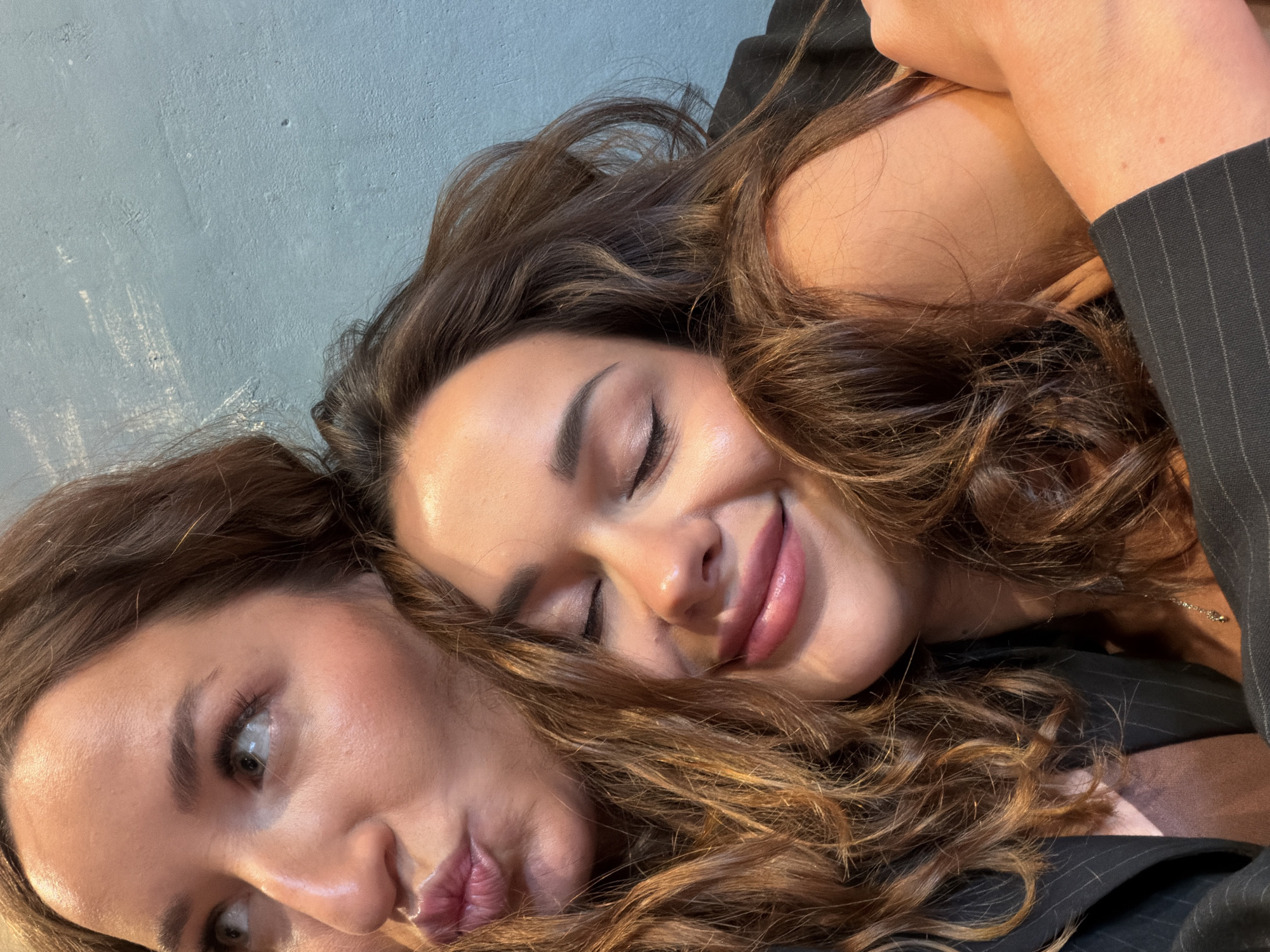 The image shows two women in close proximity, with one resting her head on the other's shoulder. The woman in the foreground has her eyes closed and is smiling, while the woman behind her is looking to the side with her lips pursed. They are both wearing dark clothing, and the background appears to be a textured blue wall. The image does not contain any specific landmarks or clues that would indicate a location in Saint Petersburg, Russia.
