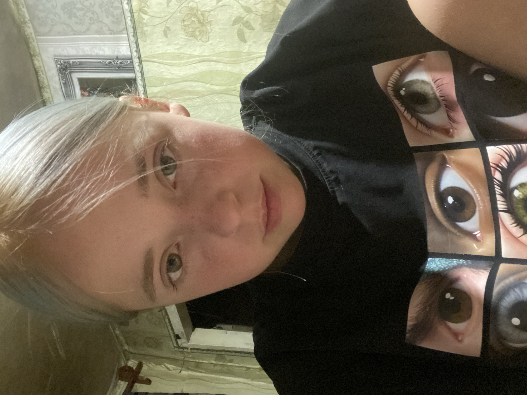 A young female with light blonde hair and freckles is looking directly at the camera. She is wearing a black t-shirt with a graphic print of several eyes. The background appears to be an indoor setting with patterned wallpaper and a doorway. There is no indication of the location being in Nova Praha, Ukraine.