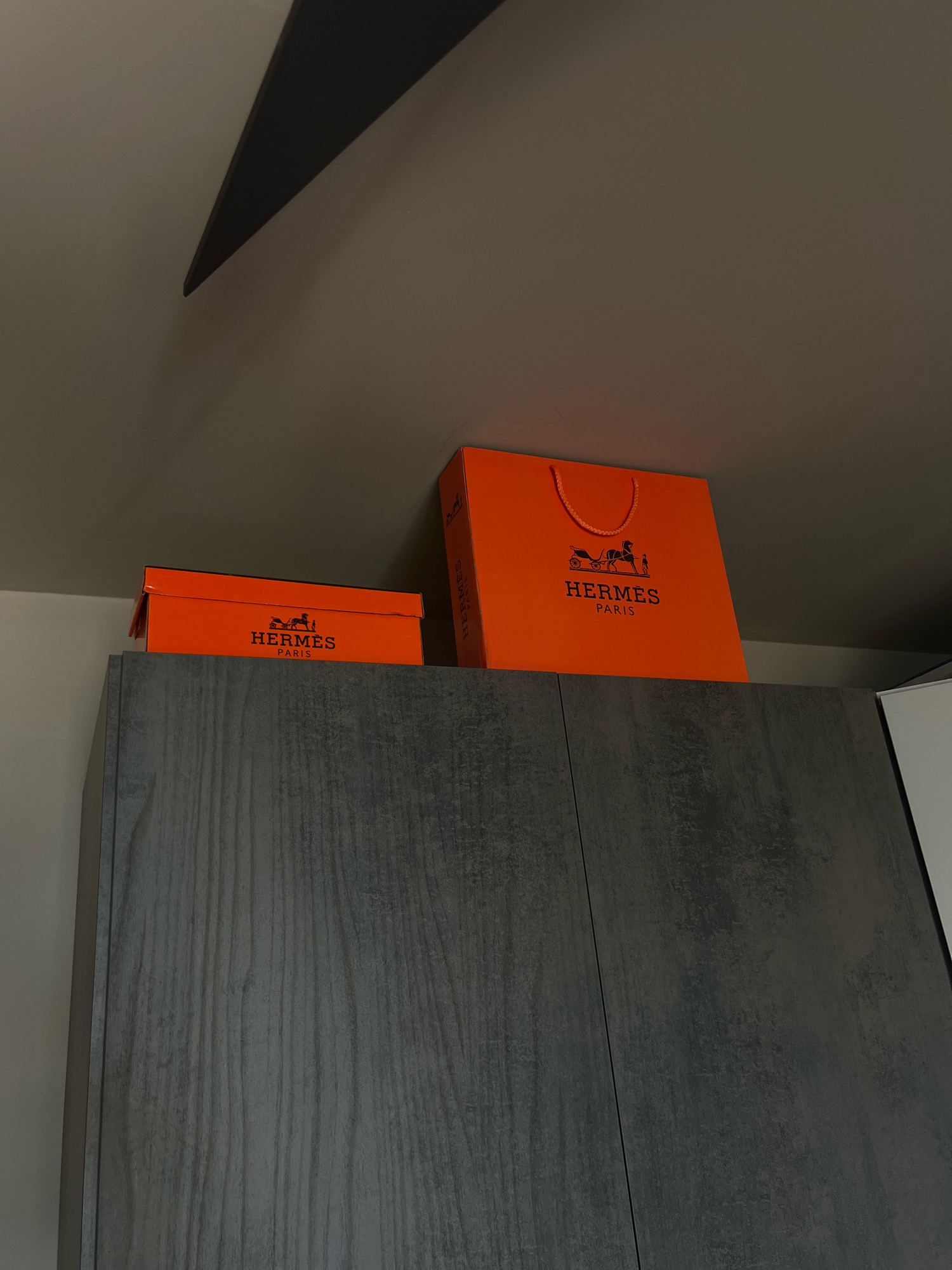 The image shows two orange Hermès boxes and a shopping bag stacked on top of dark grey cabinets. The boxes and bag are branded with the Hermès logo, which features a horse-drawn carriage. Above the cabinets and boxes, a dark diagonal shadow is cast from a ceiling fixture, possibly a fan or light. The background is a plain, neutral-colored wall. The scene appears to be an indoor domestic setting. There is no information within the image that indicates a specific location in the city of Varna, Bulgaria.