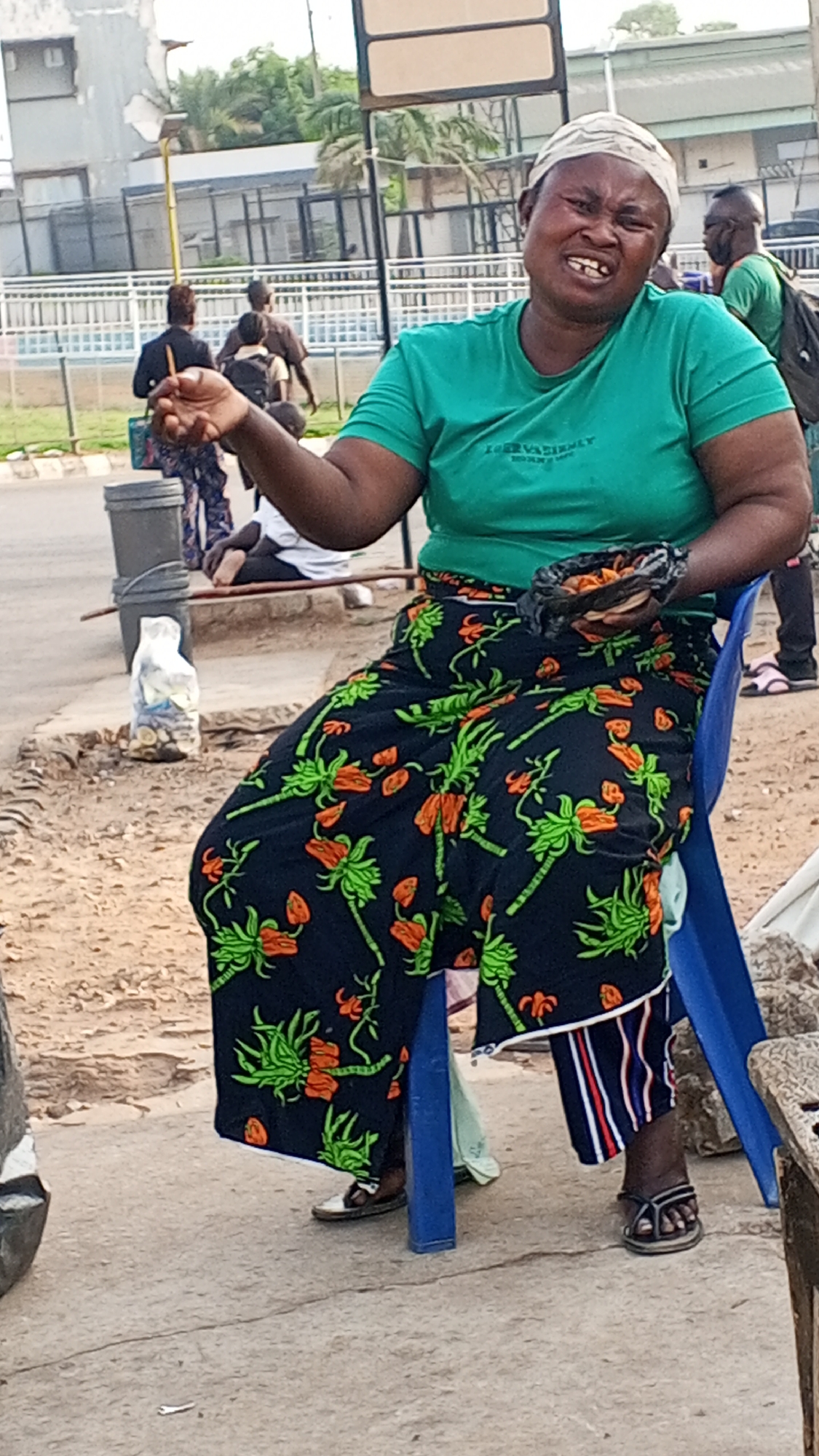 A woman sits on a blue plastic chair on a concrete surface in Maiburiji, Nigeria. She wears a green t-shirt, a black skirt patterned with orange and green floral designs, a white head covering, and sandals. She holds a handful of small, dark objects in her left hand and a bag of similar items in her right hand. Behind her, pedestrians walk along a road. There is a trash can and a white plastic bag nearby. A fence and buildings are visible in the background.