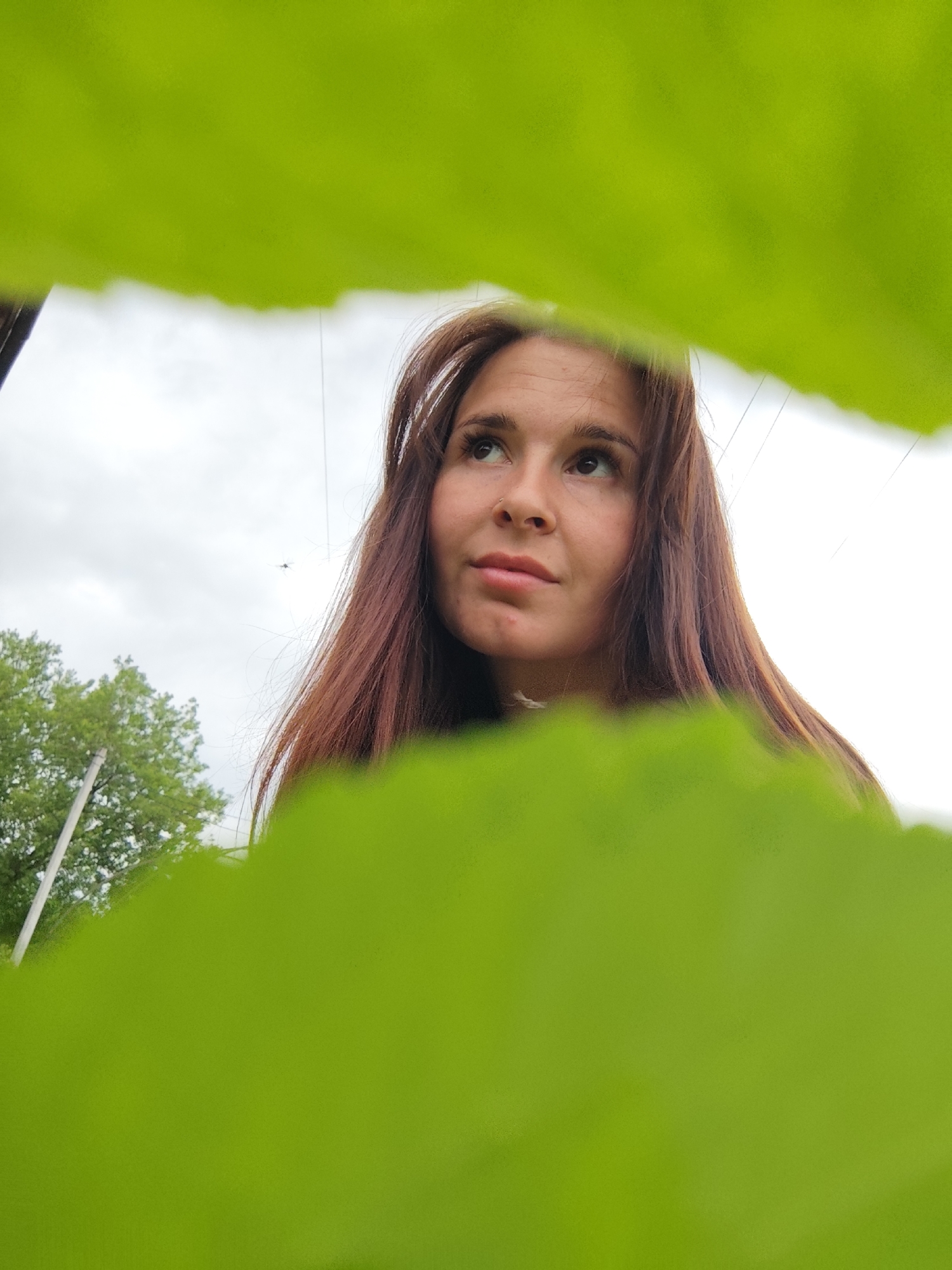 A woman with long, reddish-brown hair is partially framed by a blurry, bright green foreground. She is looking upwards and slightly to the left. The sky behind her is overcast. To the left of her, a tree and a white pole are visible. In the background, thin electrical wires can be seen against the sky. It is difficult to determine the exact location within Фоки, Russia, due to the limited visible details.