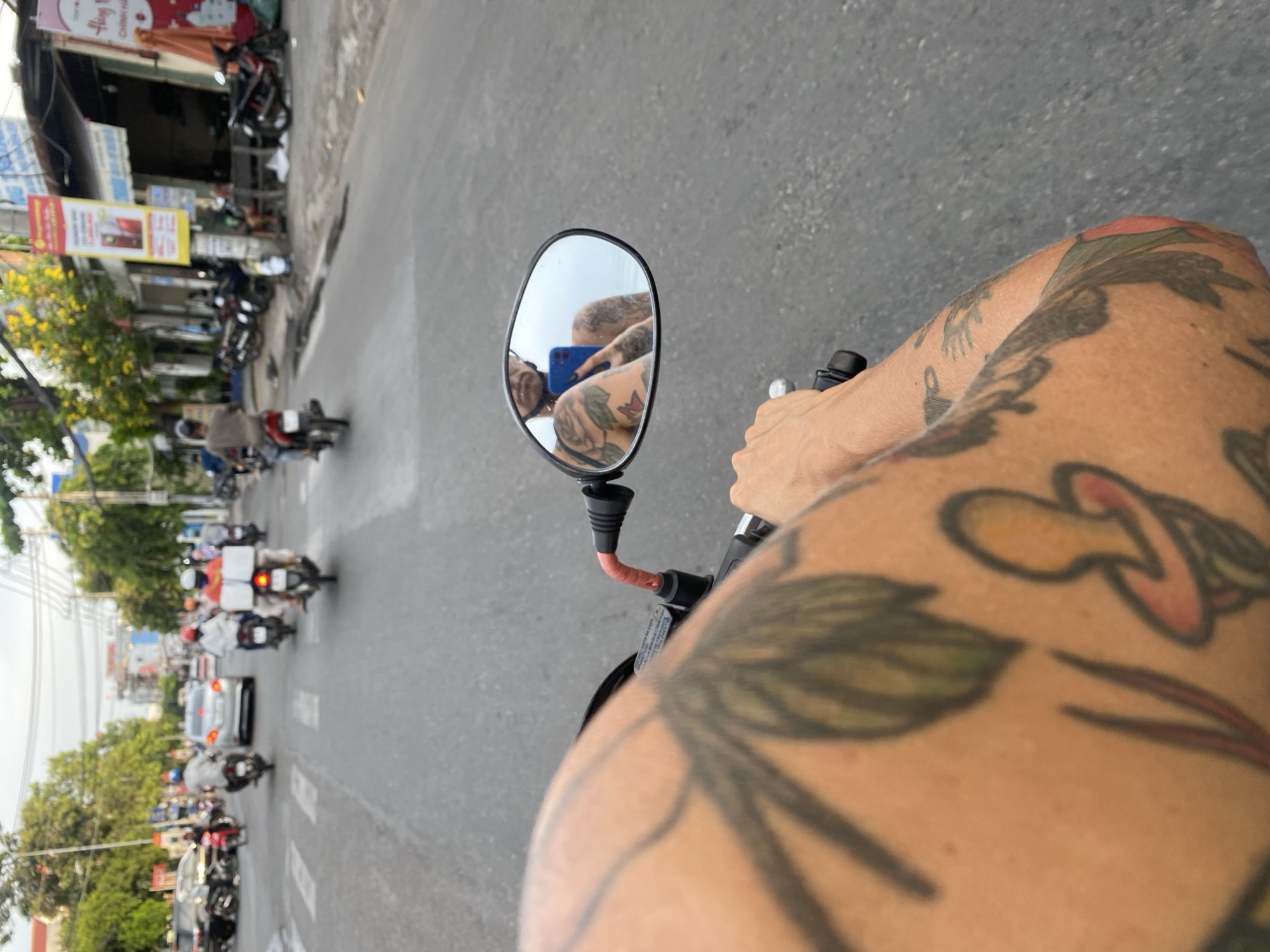 The image, taken from a first-person perspective on a moving motorcycle in Phan Thiết, Vietnam, shows the rider's heavily tattooed arm in the foreground. The side mirror of the motorcycle reflects the rider holding a blue smartphone. The background reveals a busy street with multiple motorcycles and cars traveling in the same direction. Buildings and trees line the street.