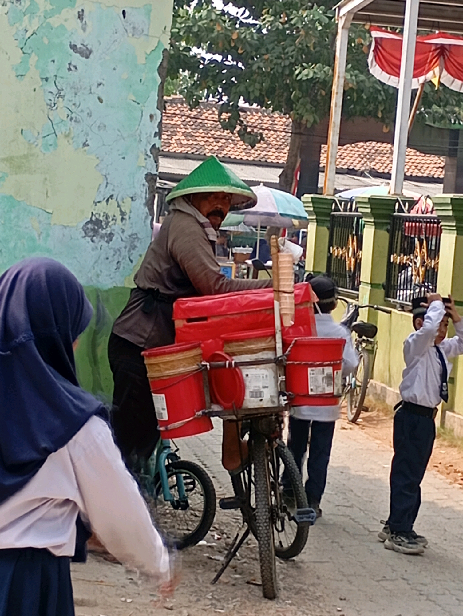 In Babelan, Indonesia, a man wearing a green conical hat and a grey long-sleeved shirt is riding a blue bicycle equipped with a large red container and two red buckets. He has a mustache and is looking to the right. To his left, a person wearing a dark blue hijab and a light-colored long-sleeved shirt is partially visible. In the background, two boys in white shirts and dark pants are standing on a dirt road. One boy is holding his hands to his head, and the other is facing away from the camera. A building with green trim and a patterned fence is behind them. The scene is outdoors, with trees and a tiled roof visible in the distance.