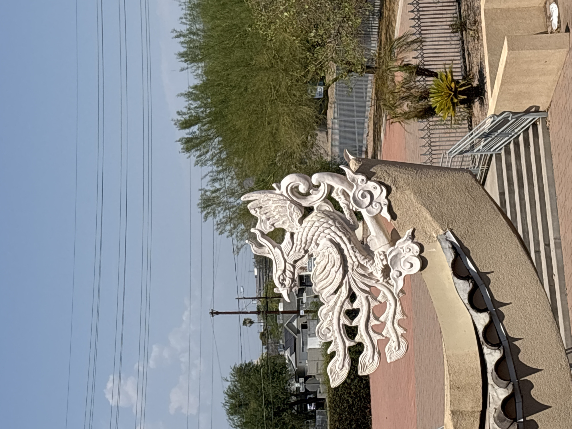 The image shows a white dragon statue on a tan structure in Phoenix, Arizona. The dragon is detailed with scales and flowing mane, perched on what appears to be the curved edge of a roof or facade, adorned with decorative tiles underneath. In the background, there are trees, buildings, power lines, and a fence. A set of stairs with a metal railing leads up towards the structure. The sky is clear and blue with a few scattered clouds. The overall setting suggests an outdoor area, possibly a temple or cultural center, under a bright, sunny day.