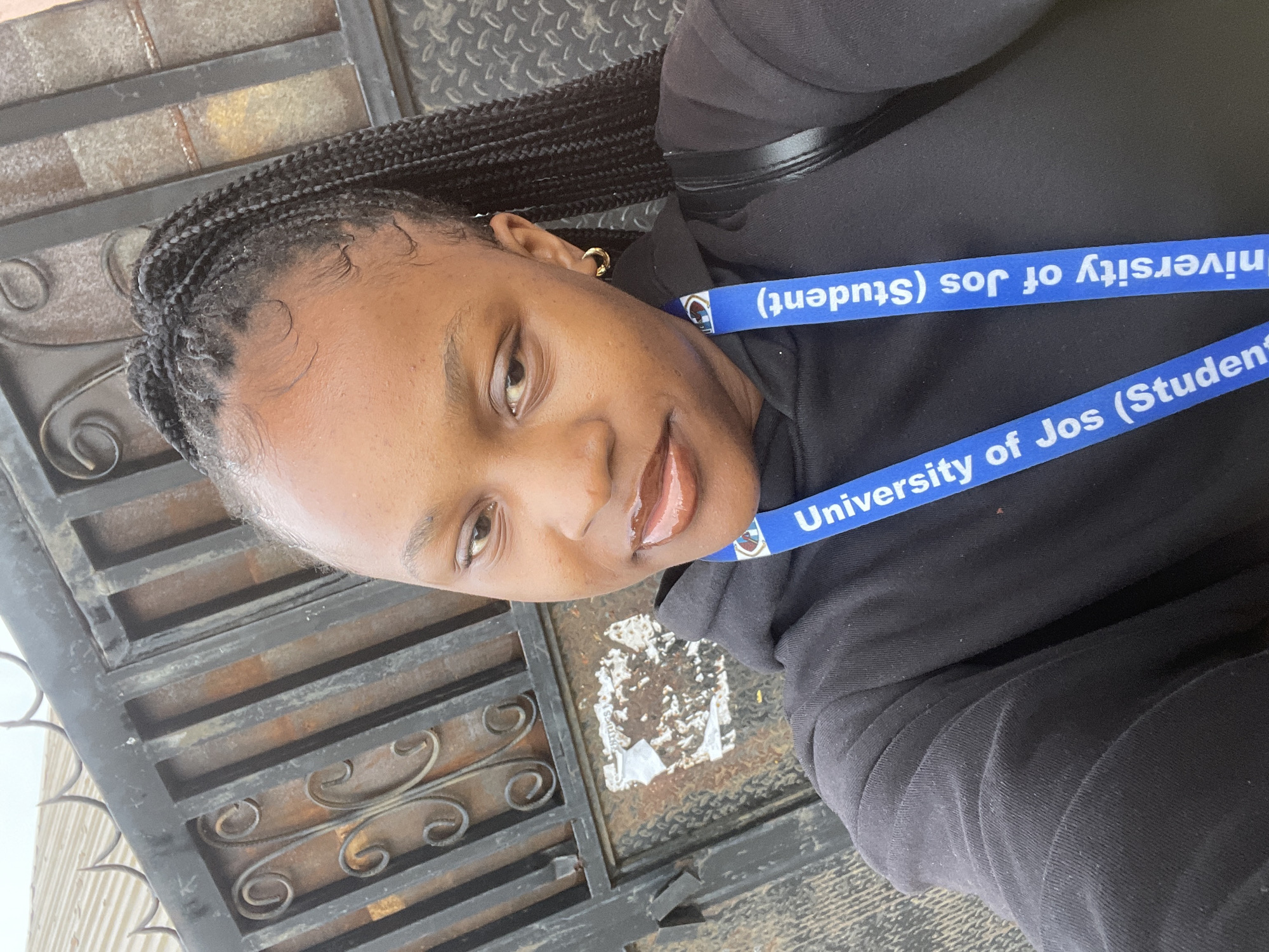 A young Black woman with dark skin and braided hair pulled back into a ponytail is taking a selfie in front of a rusty, ornate black metal gate. She is wearing a black hoodie with a blue lanyard that says "University of Jos (Student)" hanging around her neck. She has on small gold hoop earrings and appears to be wearing lip gloss. The background shows a section of the gate, which has intricate scrolling metalwork and some peeling paint or stickers on a lower panel. The ground beneath the gate is not visible. The lighting suggests it is daytime. This image was captured in Rafin Par, Nigeria.