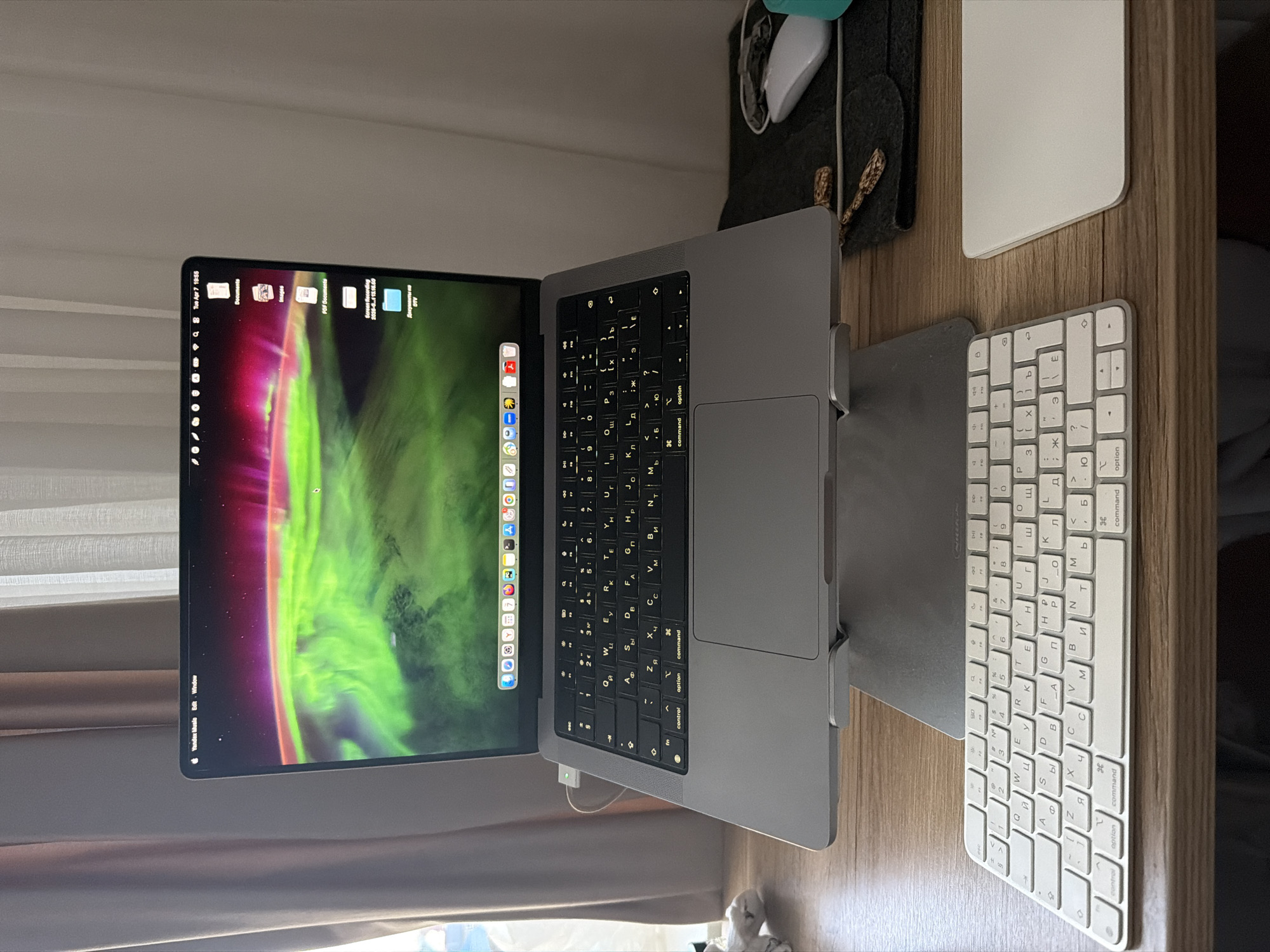 The image shows a desk setup with a MacBook Pro in a silver stand, a white Apple Magic Keyboard with Russian characters, a white Apple Magic Trackpad, and a turquoise mouse, all on a light brown wooden surface. The MacBook Pro's screen displays a green and purple abstract background and the dock has icons for applications. The time displayed on the upper left corner of the screen indicates "Tue Apr 7 19:55". A small white USB-C adapter is connected to the side of the laptop. A dark gray mat lies near the mouse. A curtain is visible in the background on the left.
There is no information in the image that allows to determine the location in the city of Chaikovsky, Russia.