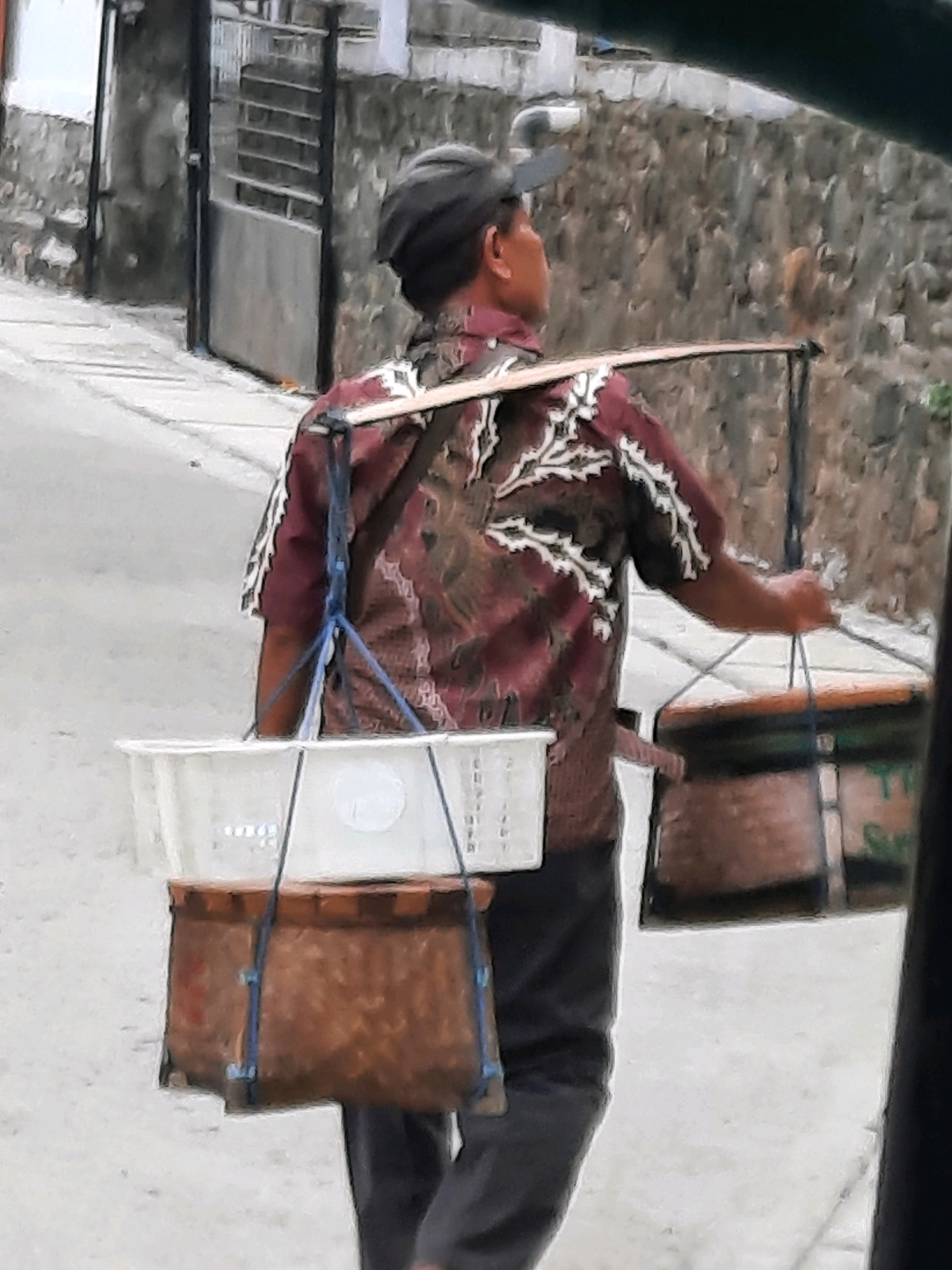 The image shows a man from the back, walking on a street in East Jakarta, Indonesia. He is wearing a dark baseball cap and a maroon shirt with a white patterned design. He is carrying a yoke across his shoulders, with two containers hanging from it. On the left, there is a white rectangular plastic container. On the right, there is a round, brown container with some green text visible. The man is wearing dark pants. The street is paved and appears to be an incline. In the background, there is a stone wall and a metal gate. The overall impression is of a street vendor.