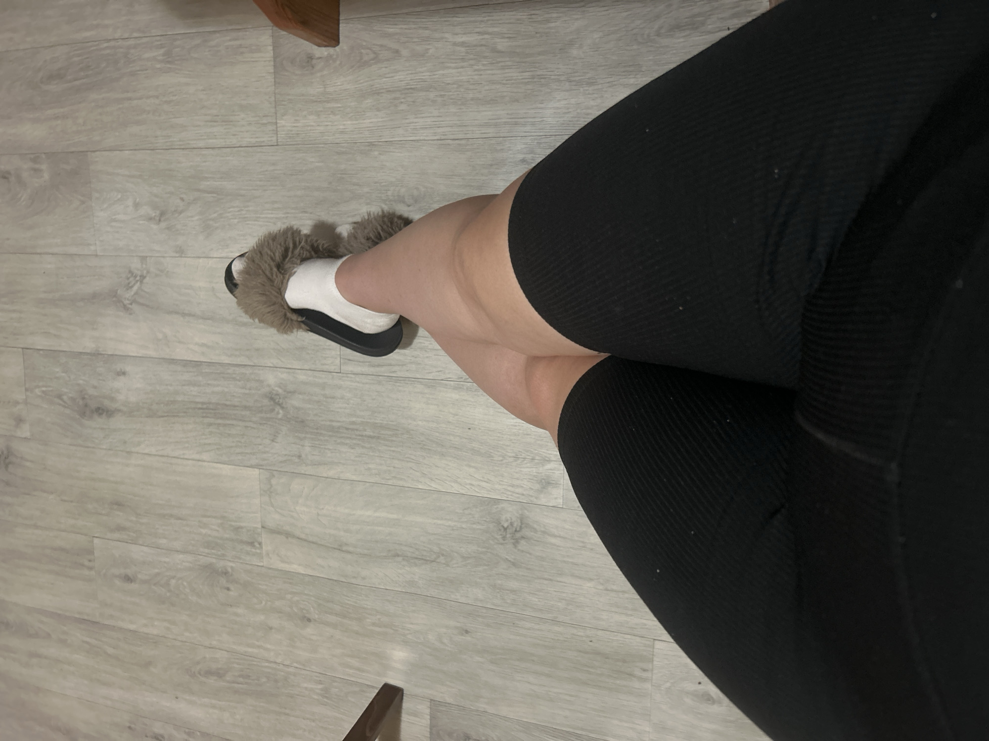 An overhead shot captures a person's legs crossed, resting on a light gray wood-patterned floor. The person is wearing black biker shorts and white socks with fuzzy brown slippers. A wooden object, possibly a chair leg, is visible in the upper left corner. The scene is indoors.